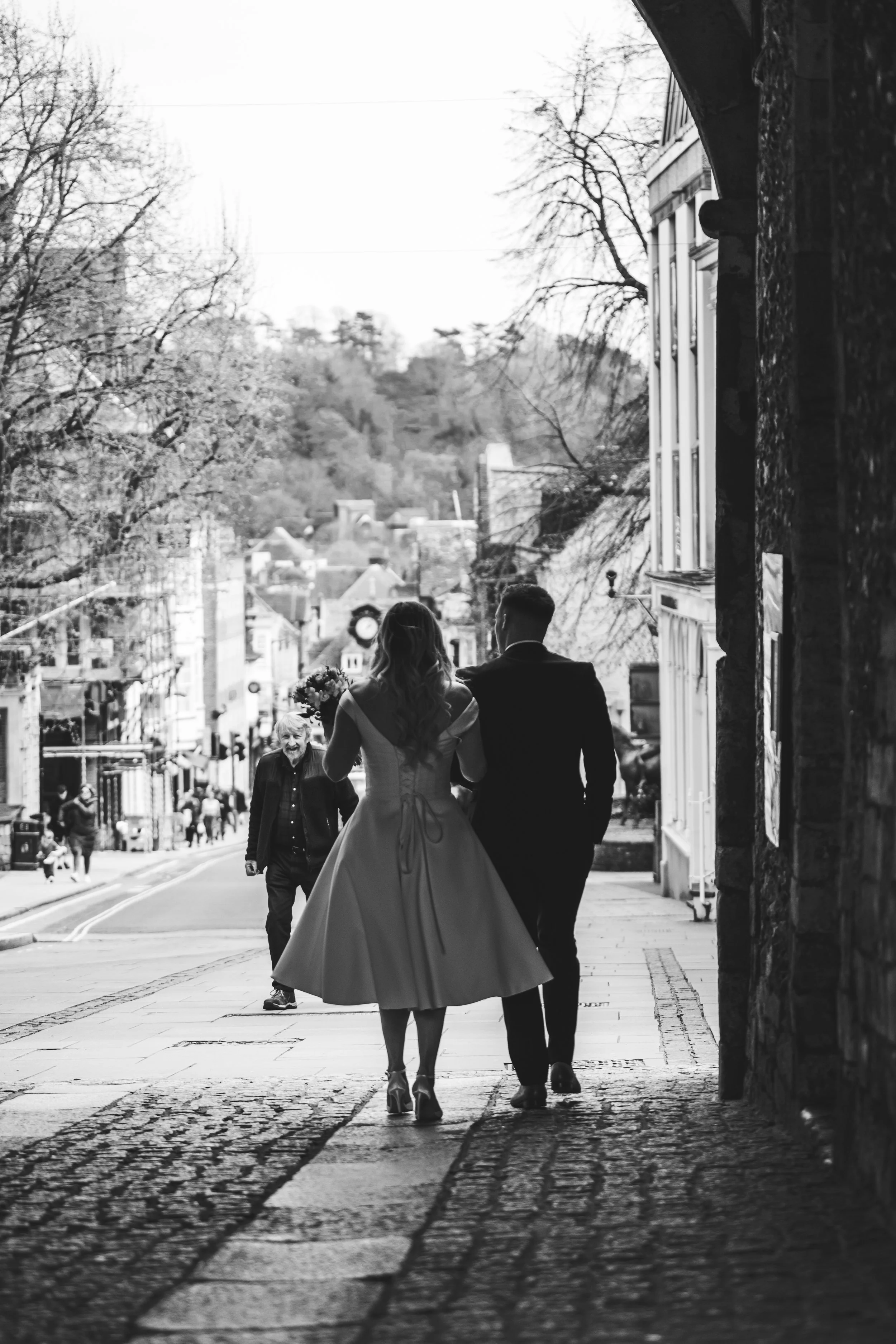 A black and white photo of a bride and groom walking hand in hand down a city street, with a woman holding flowers behind them and people in the background.