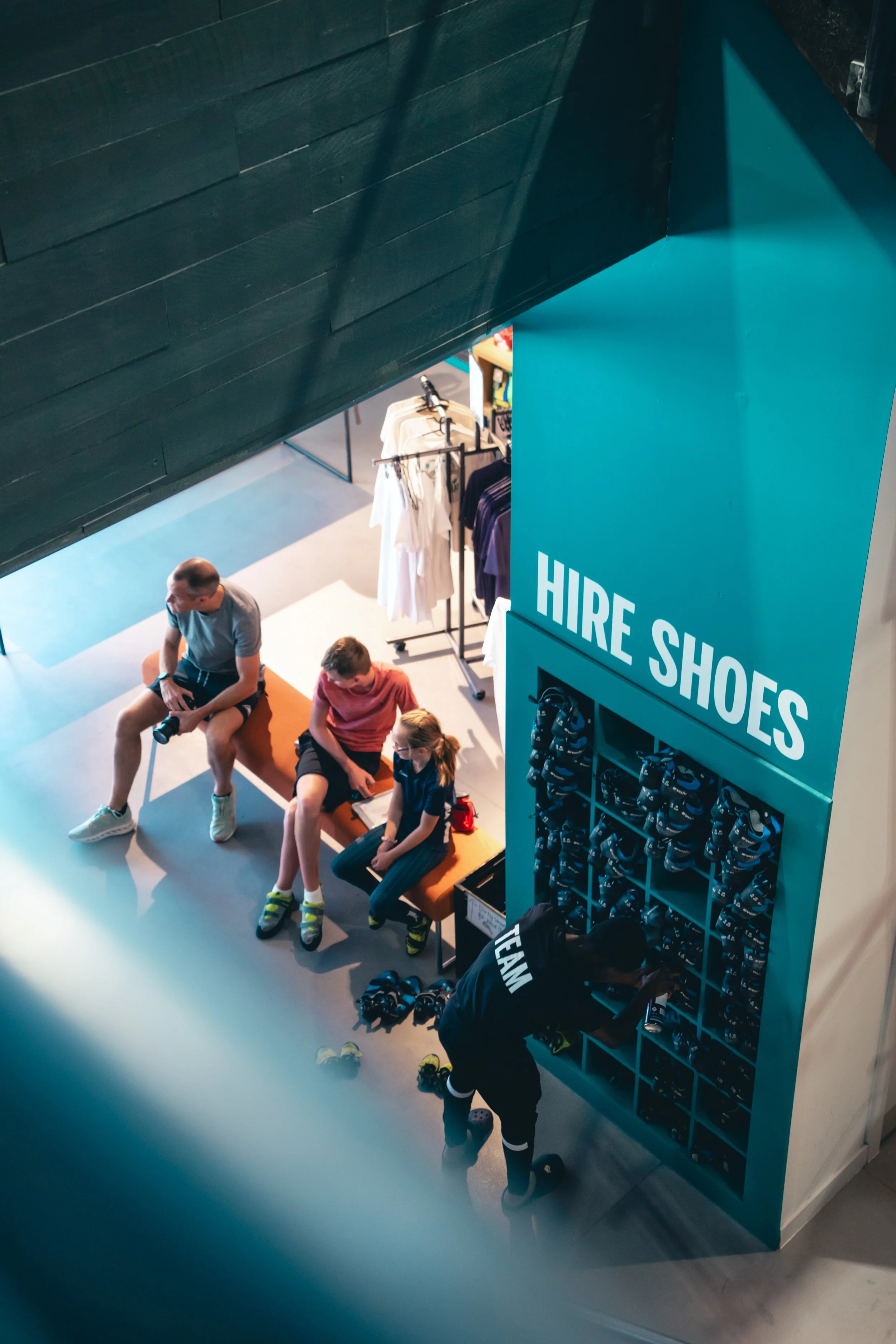 Group of people sitting on a bench near a shoe rental station in a retail store with a teal sign that says 'HIRE SHOES' and shelves of ice hockey helmets.