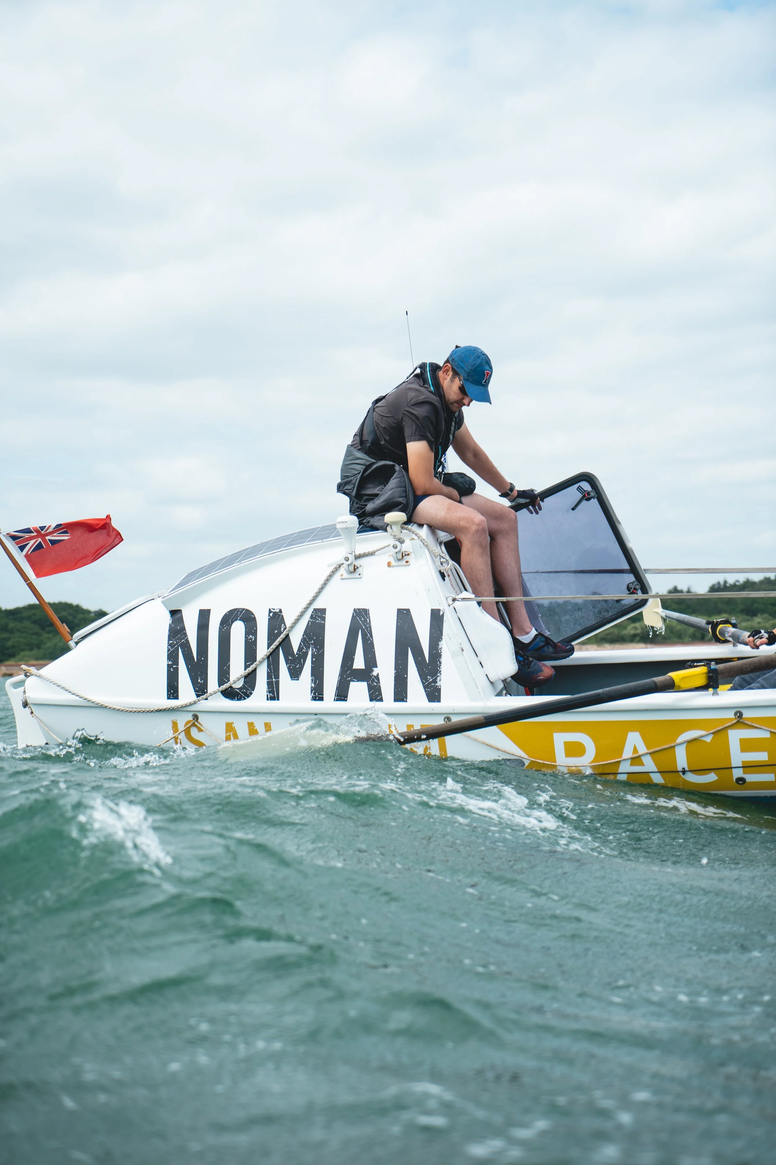 A person sitting on a small sailboat with 'NOMAN' written on the side, on open water with cloudy sky, holding a transparent windscreen, wearing a blue cap and black clothing, with a small red flag at the stern.