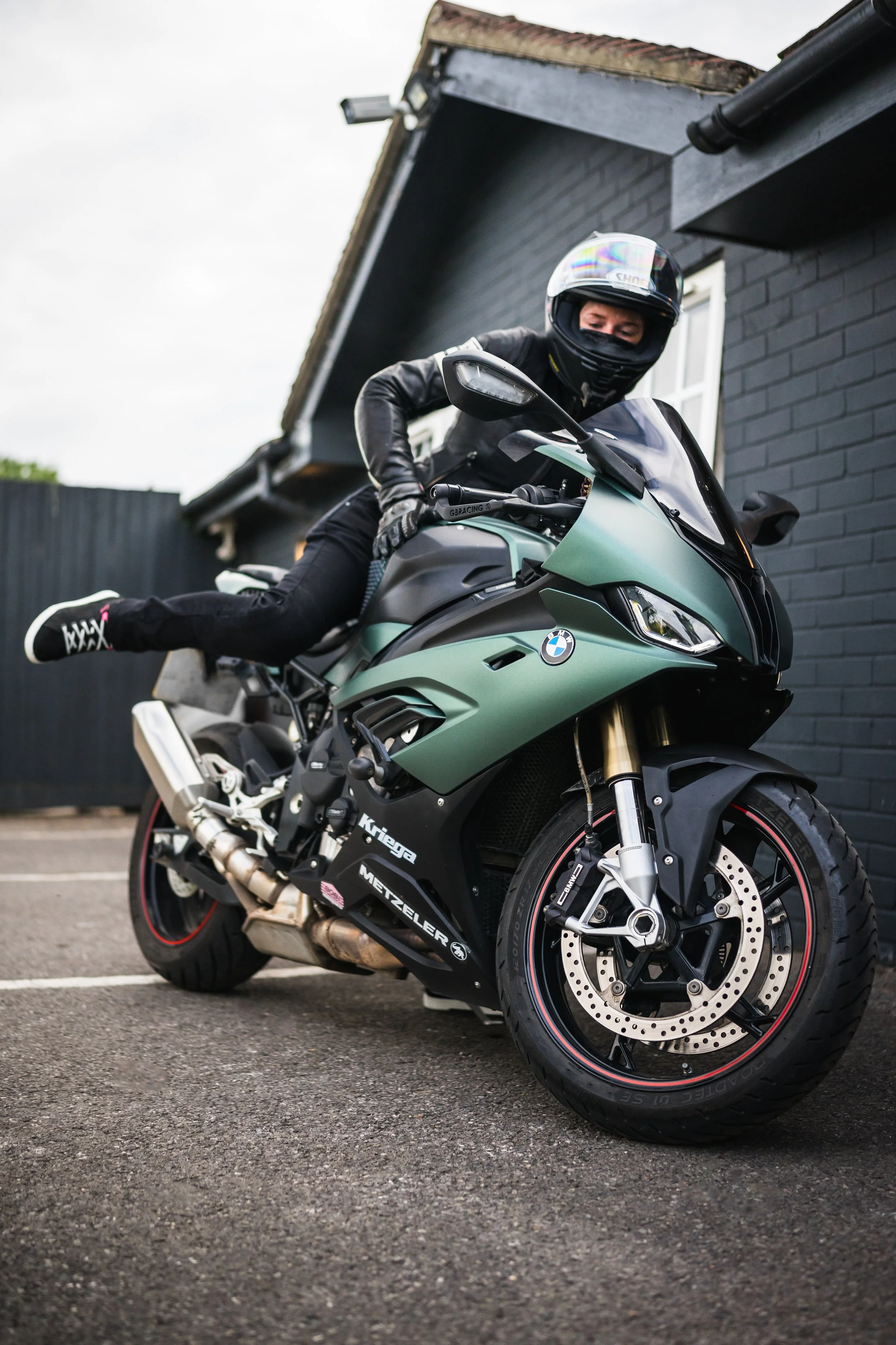 A person wearing a helmet and black leather jacket riding a green BMW motorcycle with black and red accents, parked on a paved surface near a black brick building.