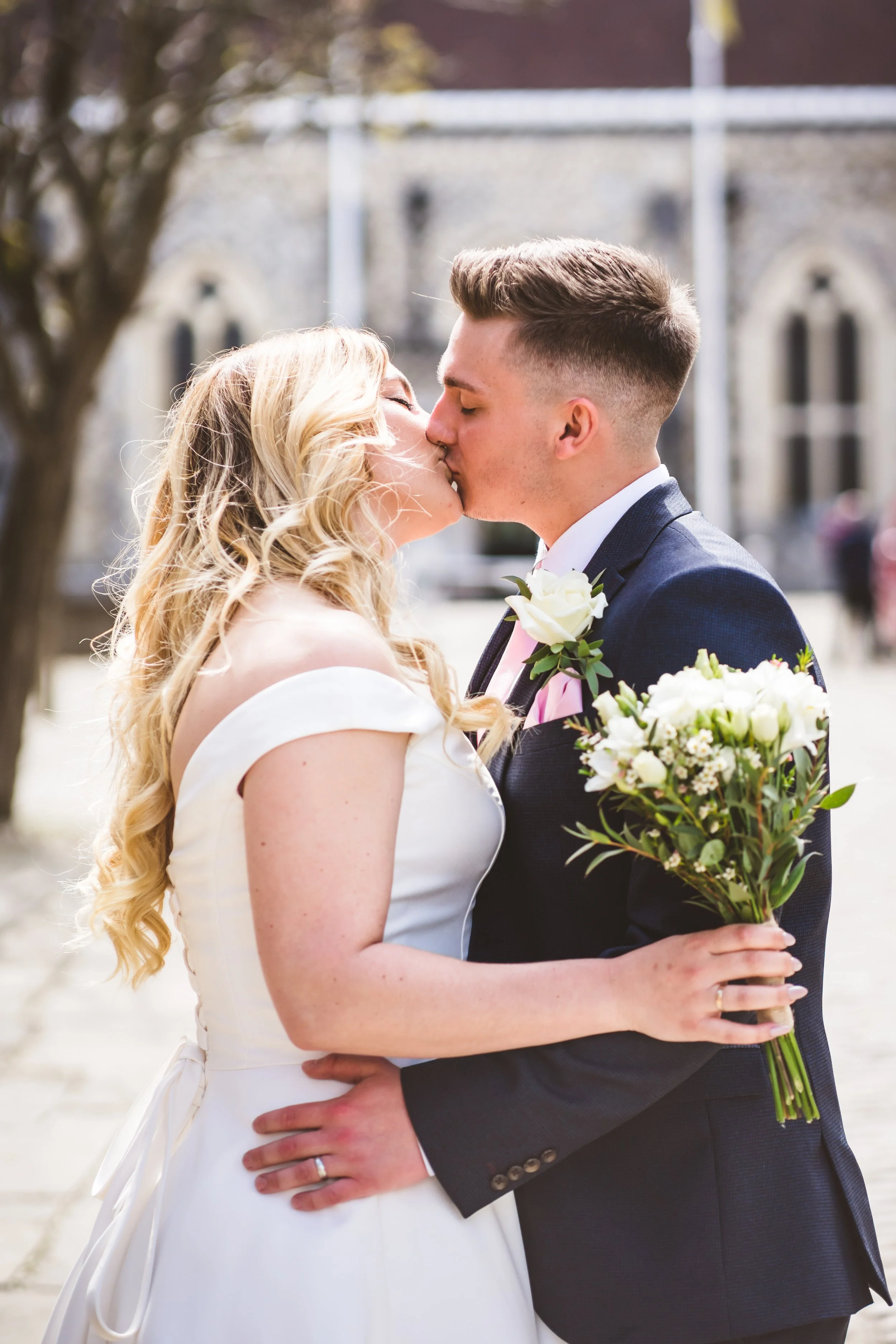A newlywed couple sharing a kiss outdoors, with the bride in a white off-the-shoulder dress and the groom in a dark suit holding a bouquet of white flowers.