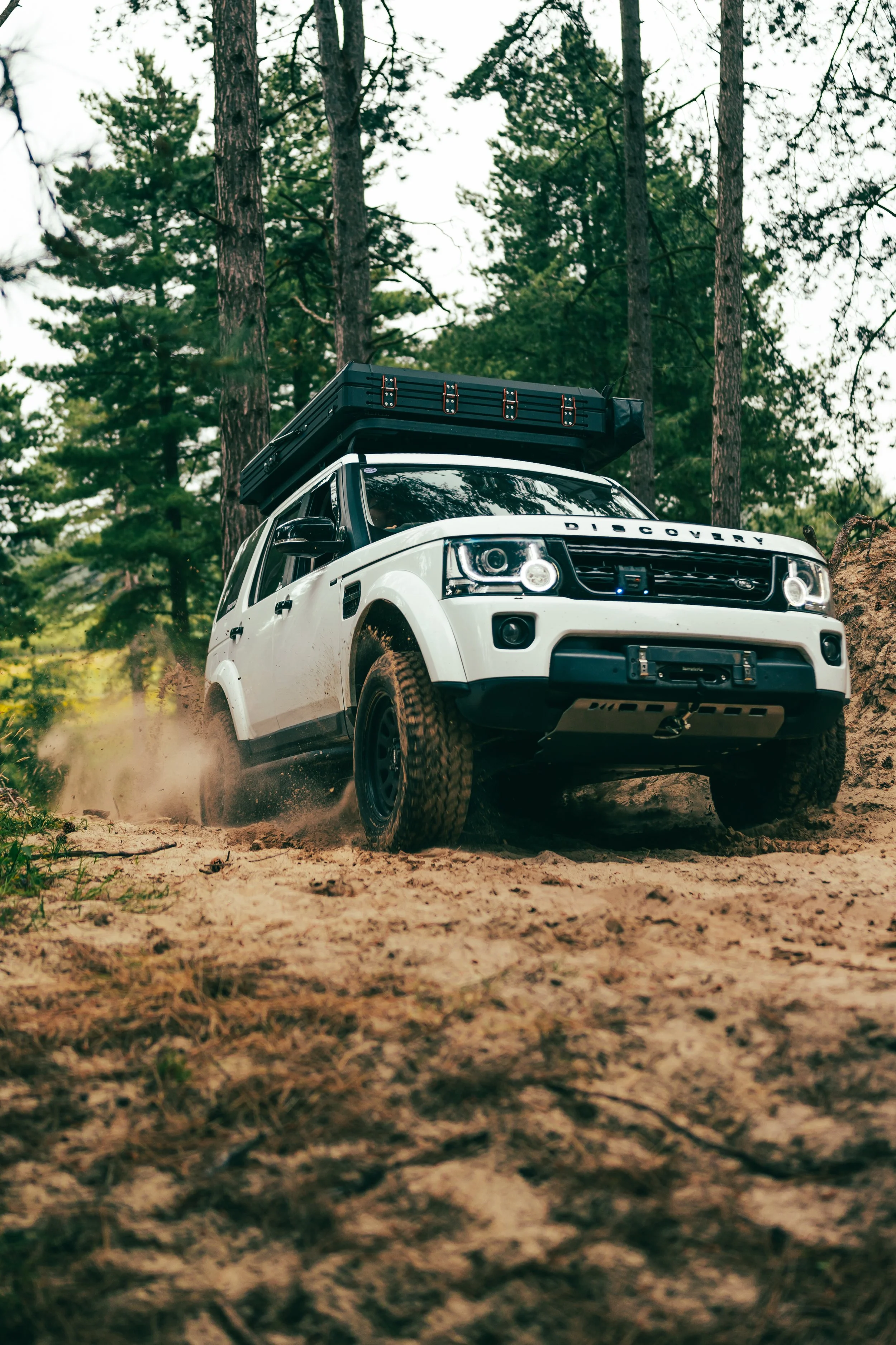 A white off-road vehicle driving through a dirt trail in a forest with green trees, kicking up dust behind it.