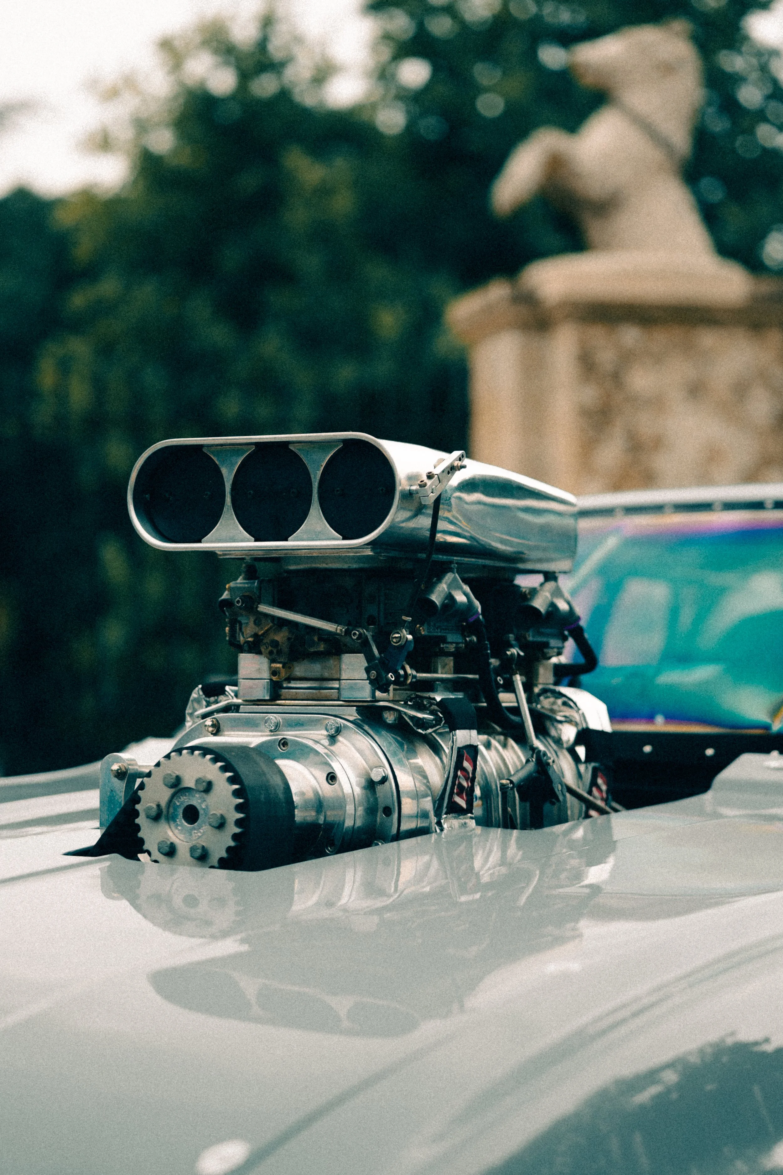 Close-up of a car engine with a supercharger installed on the hood, with a blurred background of trees and a stone horse statue.