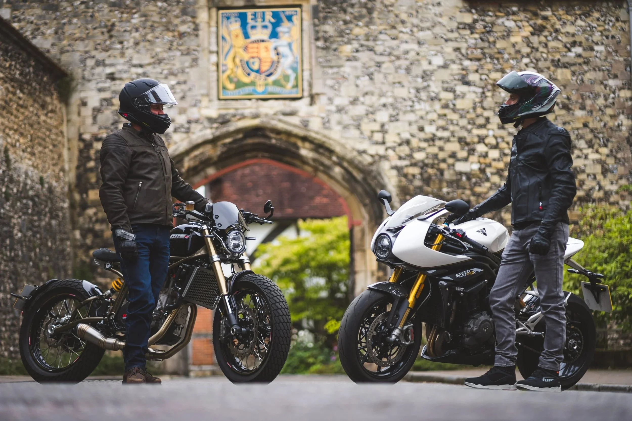 Two motorcyclists wearing helmets and jackets face each other with their motorcycles, standing in front of an old stone archway with a coat of arms above it.