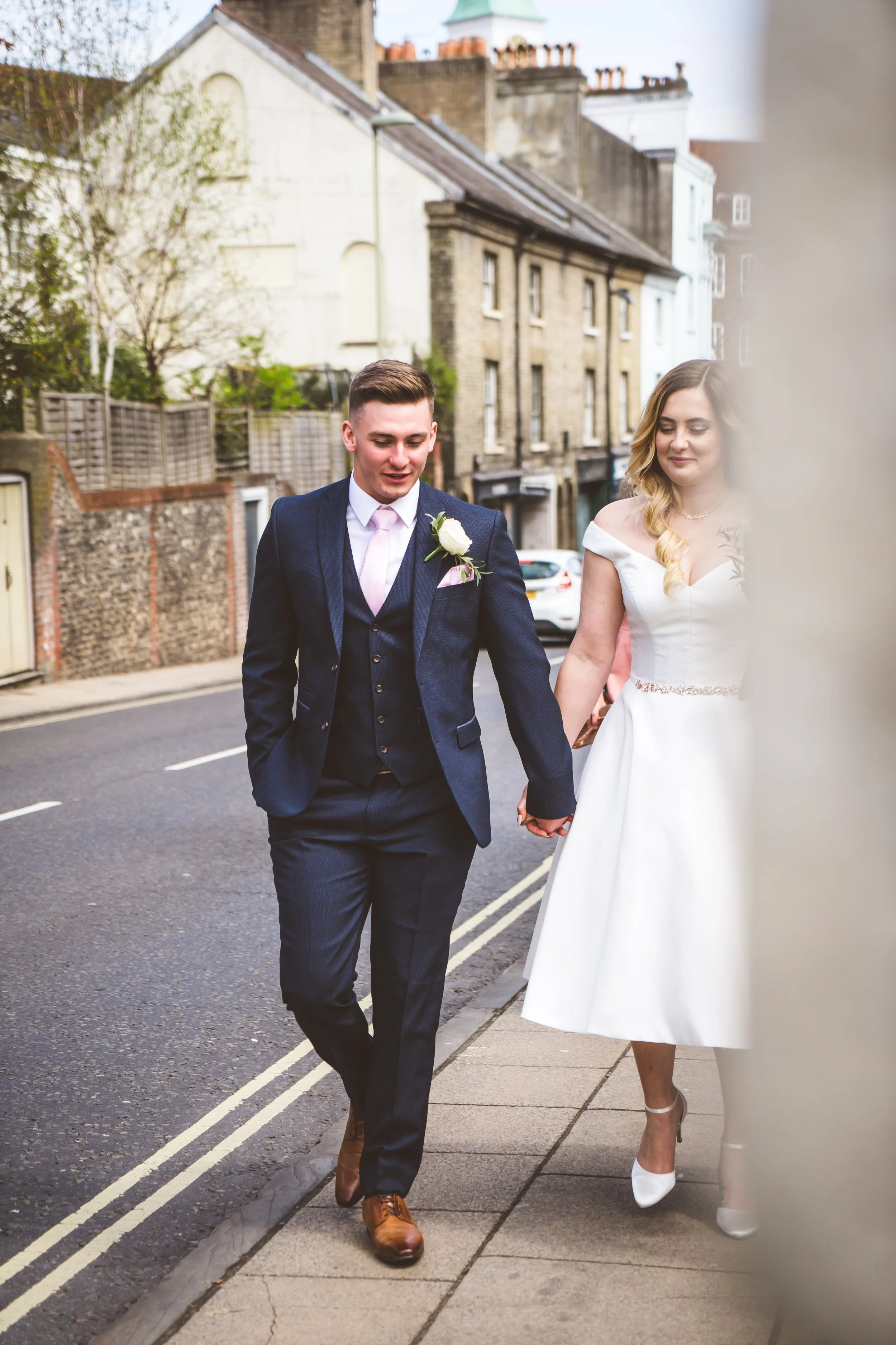 A bride and groom holding hands and walking on a street in wedding attire, with the bride wearing a white dress and the groom in a navy suit, surrounded by residential buildings.