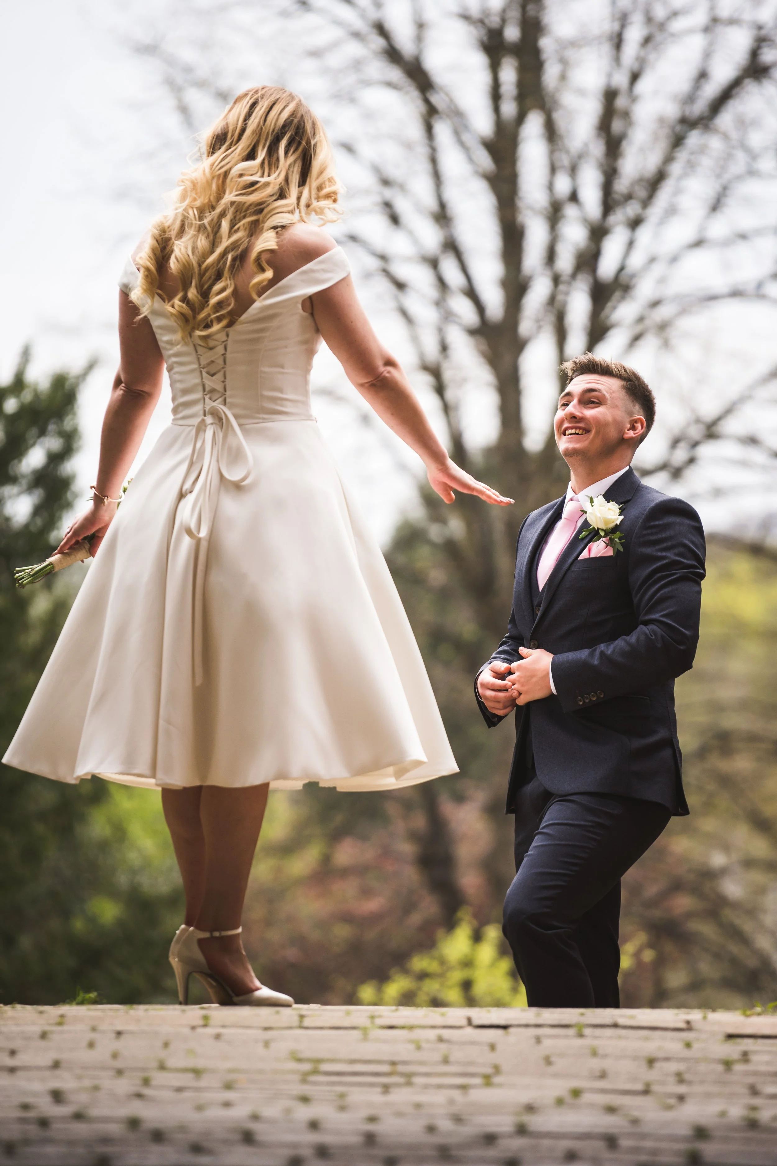 A woman in a white dress standing on a ledge, reaching towards a smiling man in a suit, outdoors with trees in the background.