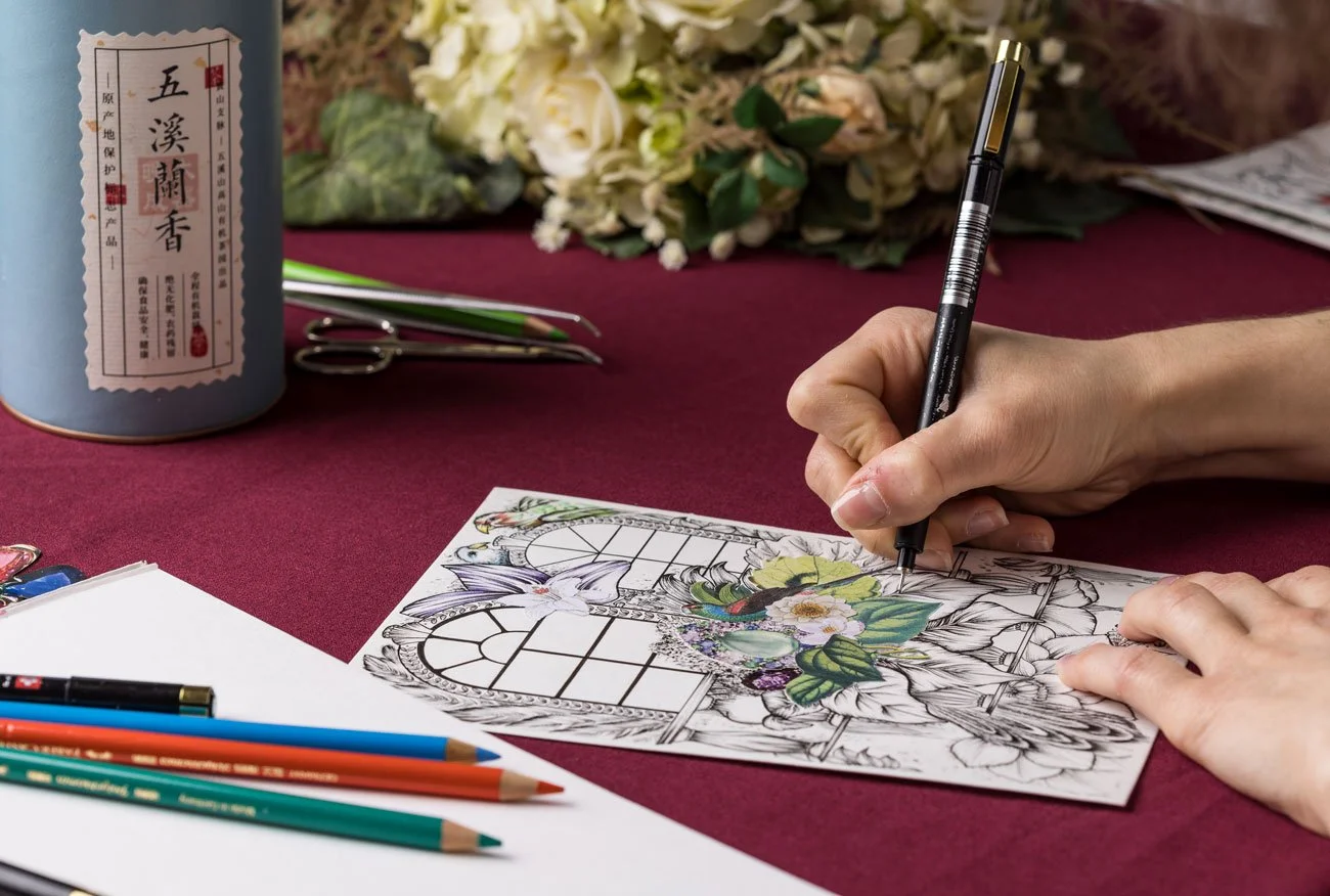 Une personne colorie un dessin détaillé de fleurs et de papillons avec des crayons et un stylo, sur une table rouge. Il y a des fleurs fraîches, des ciseaux, un pinceau, un pinceau à huile, et un grand tube d'encens en boîte à côté.