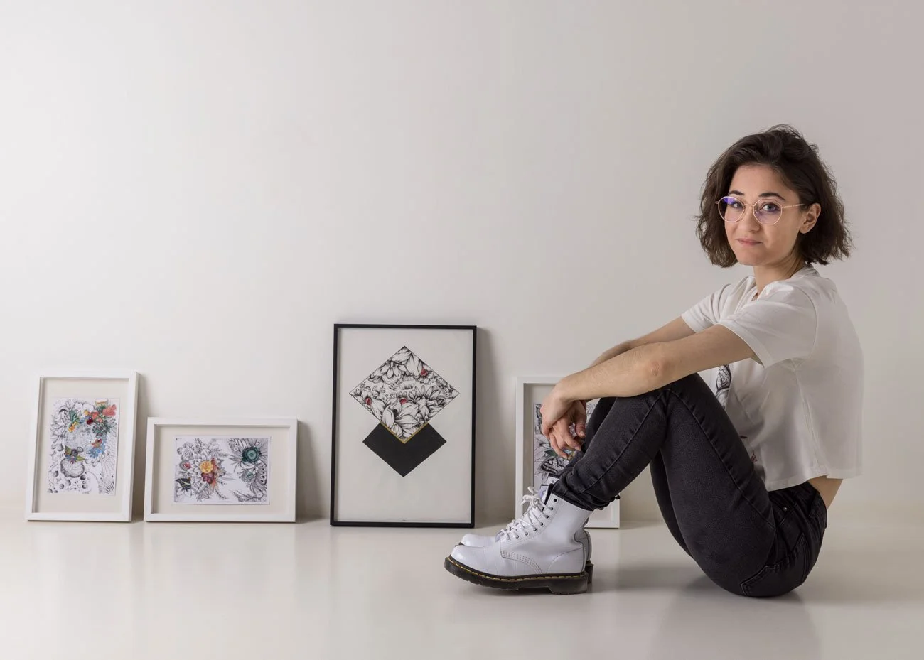 Jeune femme assise sur le sol, portant des bottes blanches, lunettes, vêtue d'un t-shirt blanc et d'un jean noir, à côté de quatre cadres avec des dessins ou des œuvres d'art sur un mur blanc.