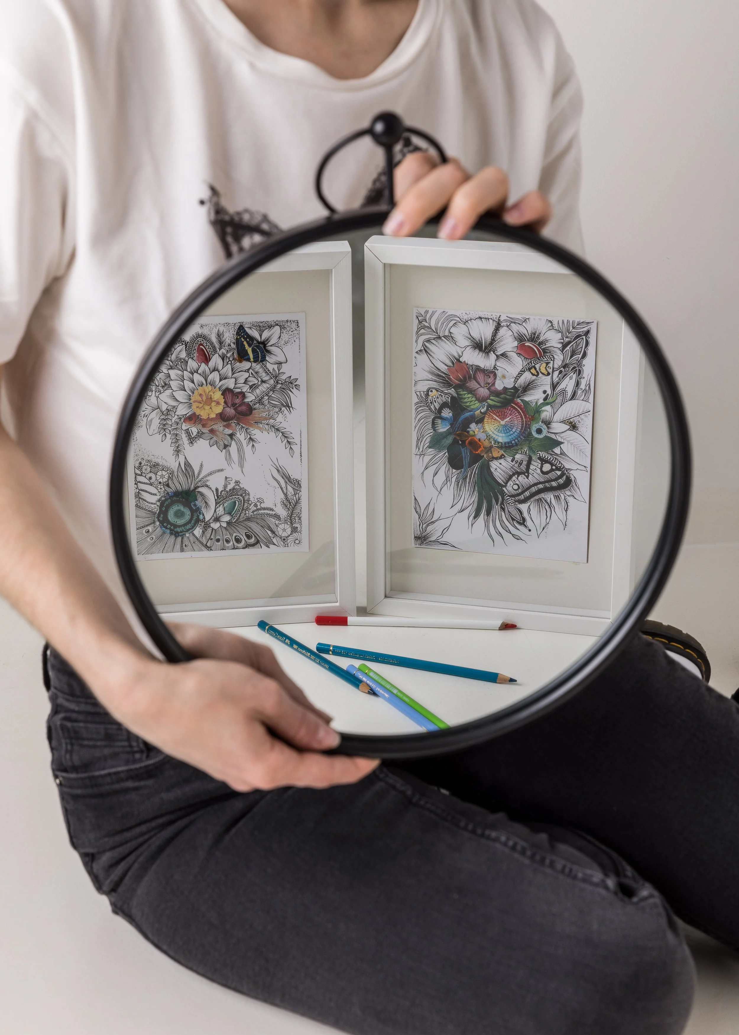 Une personne tient une lampe de poche devant un panneau avec des dessins en noir et blanc de fleurs, d'insectes et d'oiseaux. Les dessins sont colorés de façon partielle avec des crayons de couleur. La personne porte un t-shirt blanc avec un dessin noir et un pantalon gris.