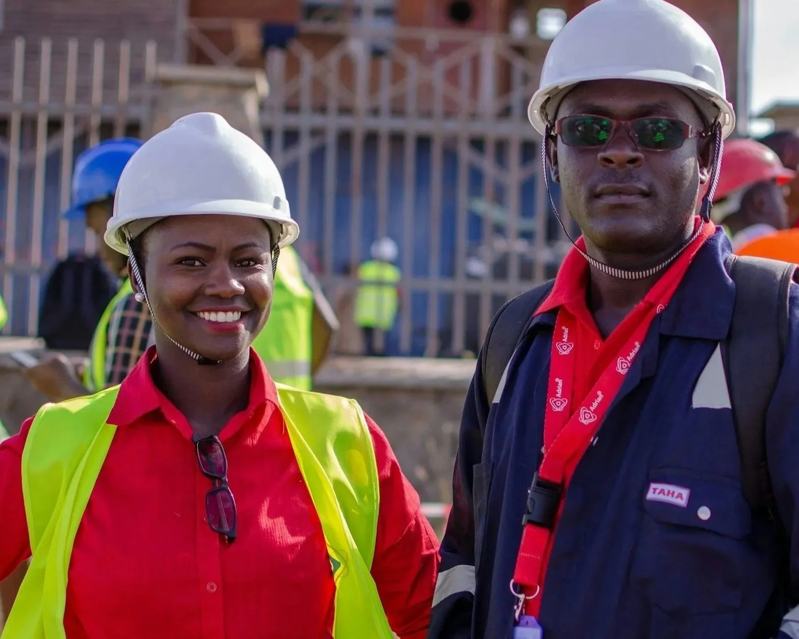 Two construction workers wearing hard hats and safety vests at a construction site. The woman has a bright smile, and the man is wearing sunglasses and a serious expression.