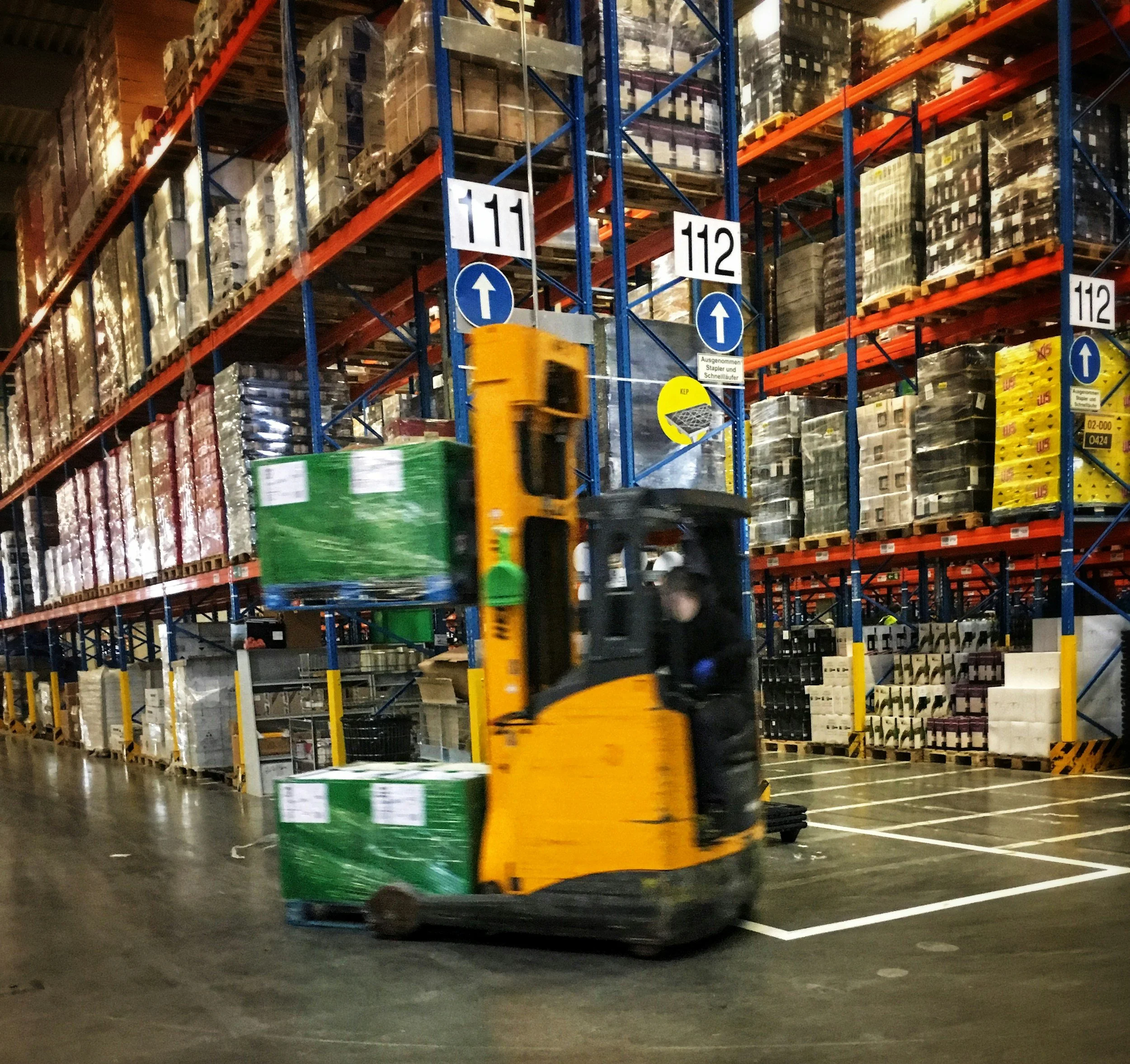 A forklift moving a pallet wrapped in green plastic inside a large warehouse with tall shelves filled with boxes and pallets.