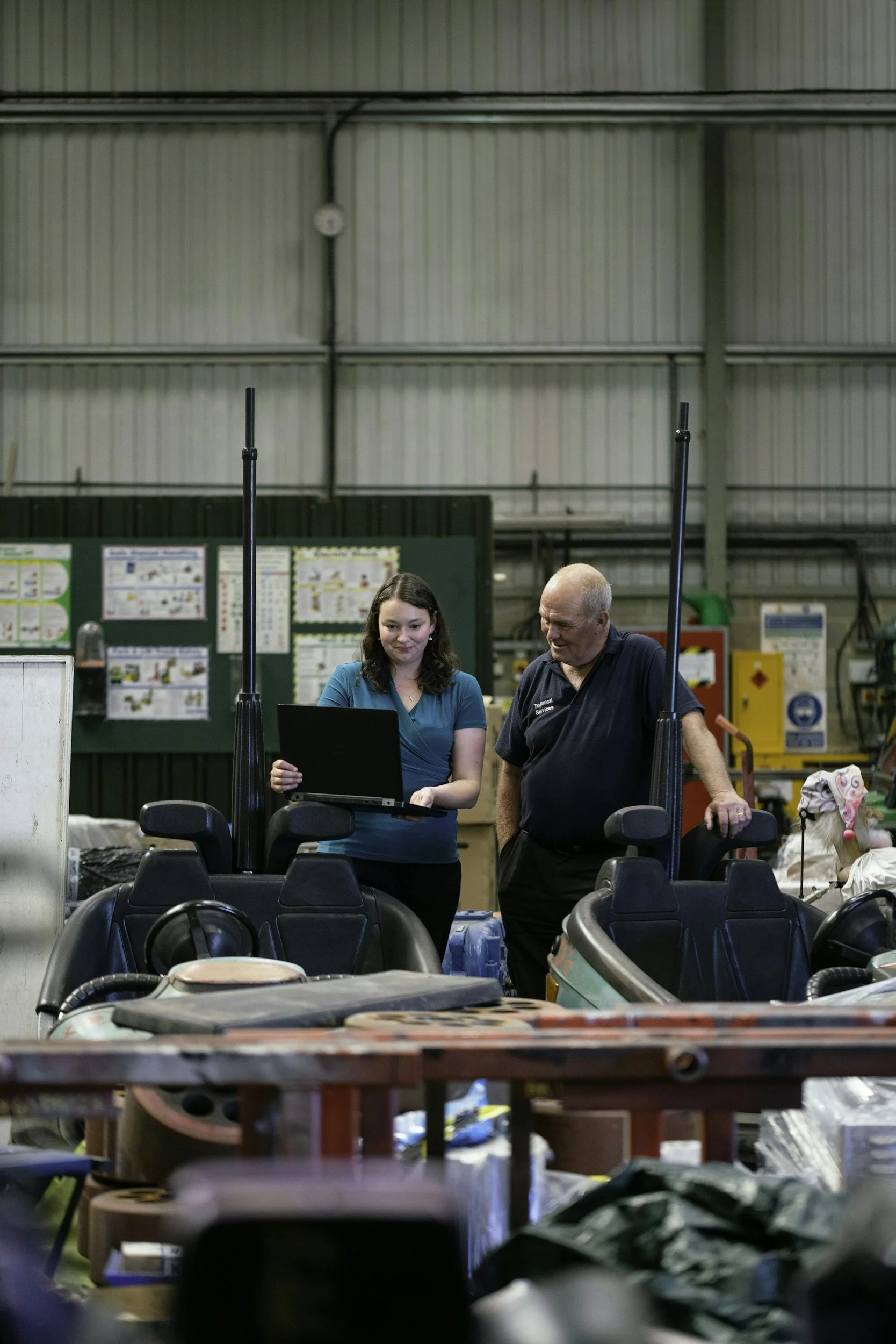 A woman and a man stand together in a large industrial space, looking at a laptop. The woman holds the laptop and smiles, while the man looks at the screen with a curious expression. There are two chairs in front of them and various equipment and supplies surrounding them.