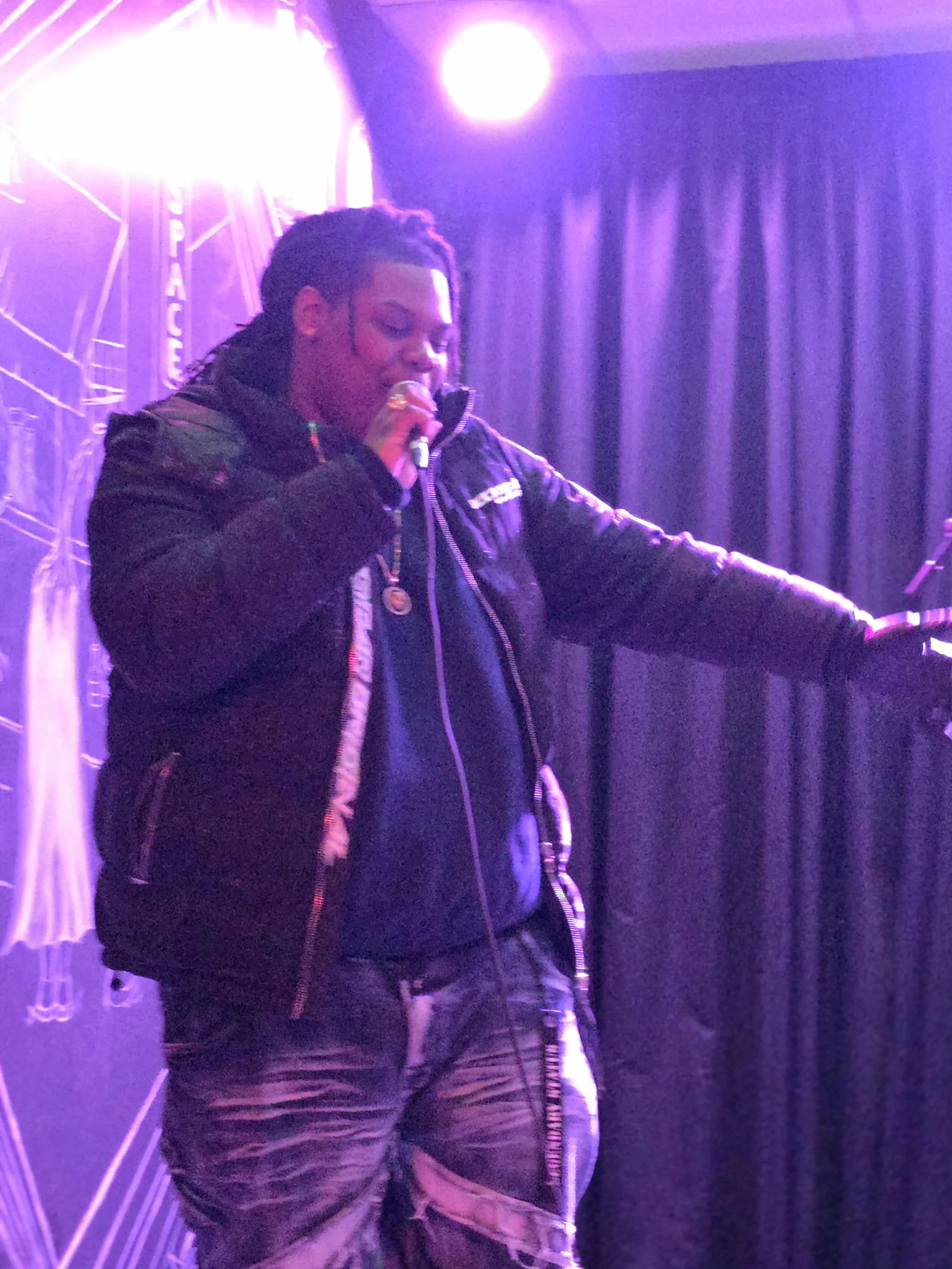 Person performing on stage with purple lighting, wearing a black jacket and camo pants, holding a microphone.