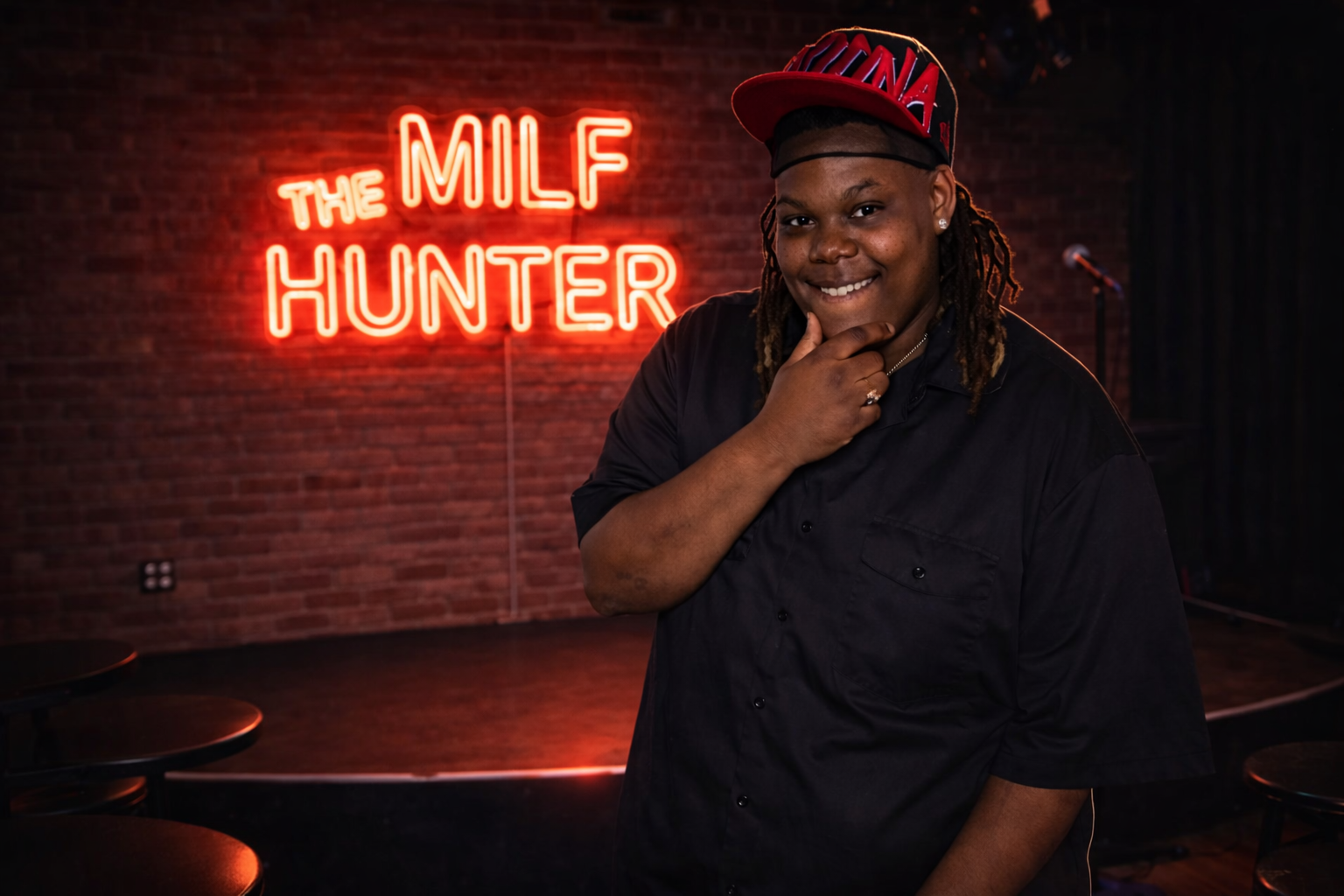 A woman with dreadlocks wearing a black chef's coat and a red cap, smiling and touching her chin, standing in front of a red neon sign that reads 'The MILF HUNTER' on a brick wall.