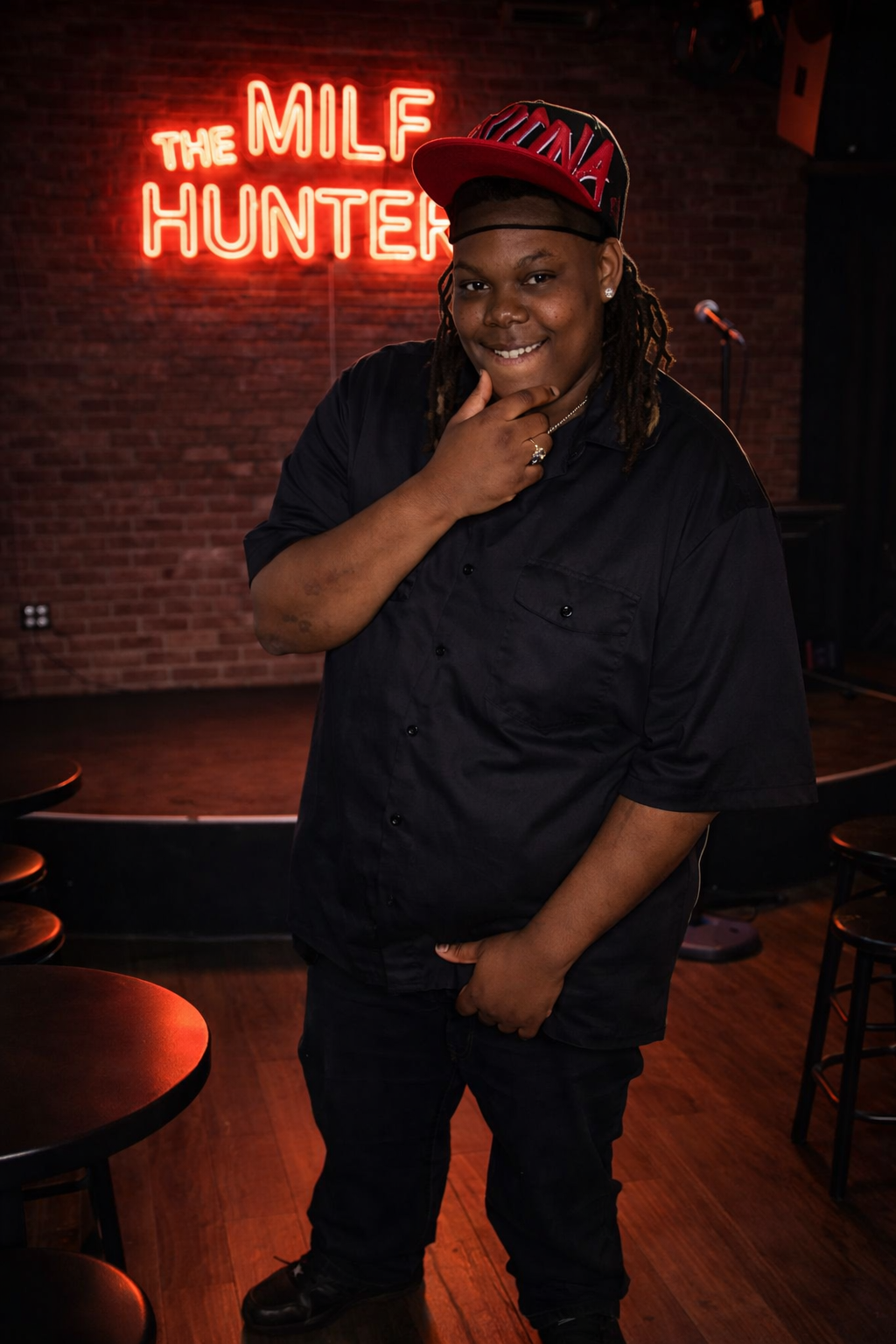 A woman with dreadlocks wearing a black shirt, red baseball cap, and jewelry, posing with a smile in a comedy club with a brick wall and neon sign that reads 'The MILF Hunter' in the background.