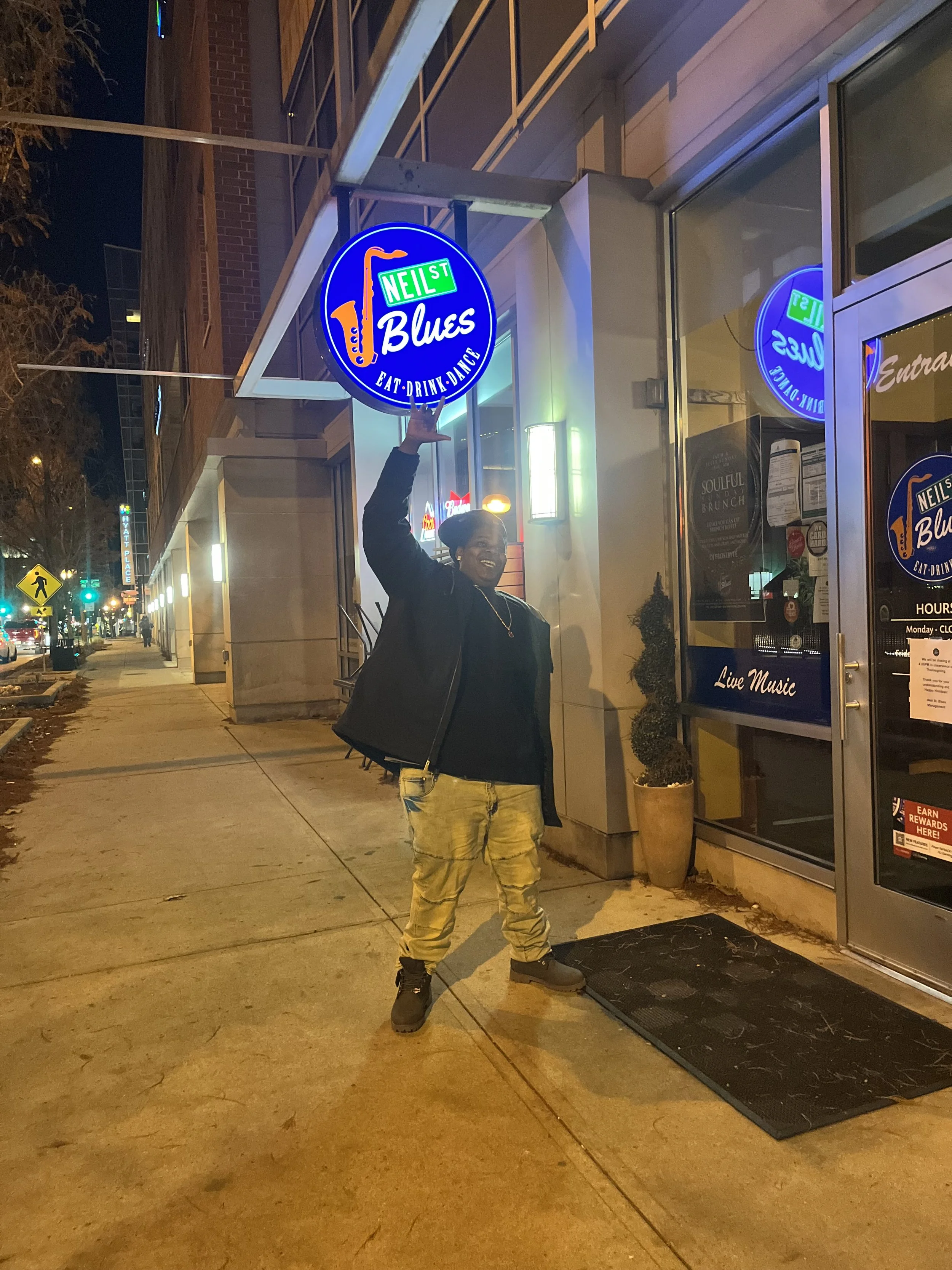 A person standing on a sidewalk at night, holding a neon sign for Neil Street Blues restaurant. The sign is circular, blue, with a saxophone graphic, and text that says 'Neil St. Blues Eat-Drink-Dance.' The person is smiling, wearing a black jacket, black shirt, yellow camouflage pants, and tan boots.