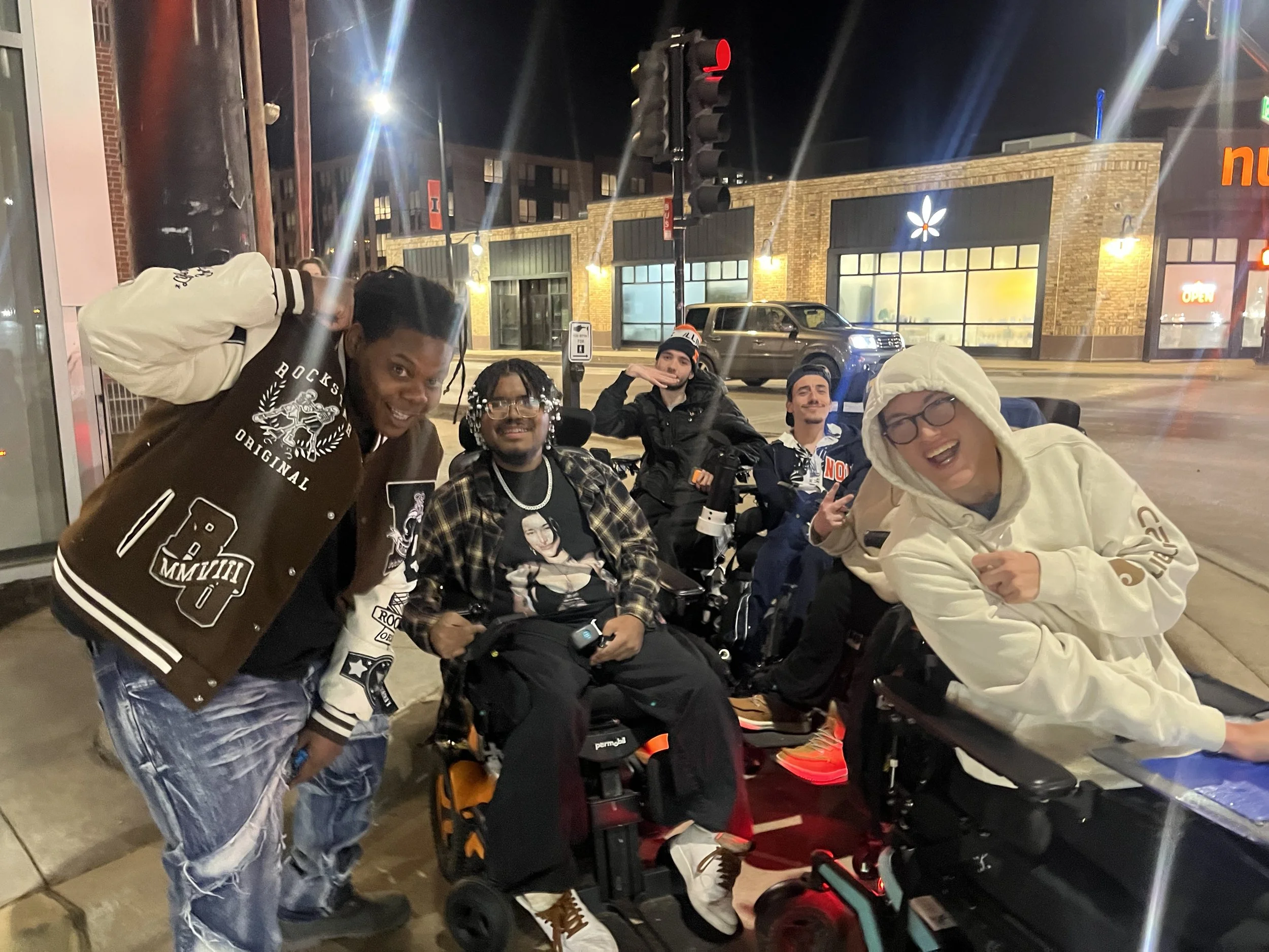 A group of five diverse young adults posing cheerfully on a city street at night, with some in wheelchairs and some standing, near a storefront with illuminated windows.
