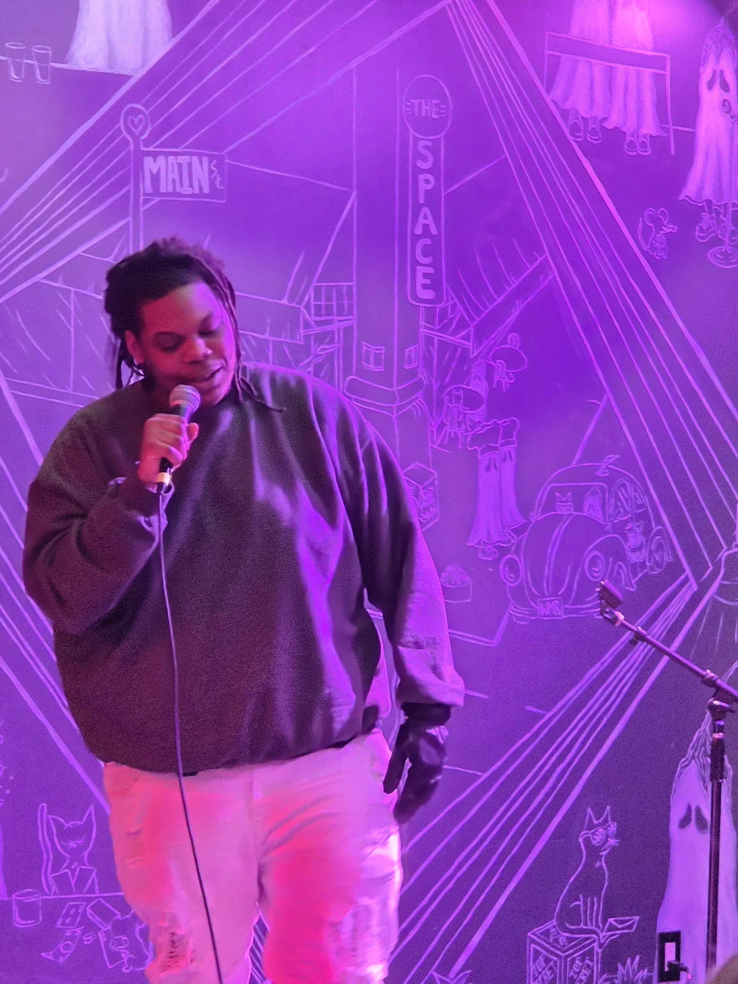 Person with dreadlocks holding a microphone, standing in front of a vibrant purple neon mural of a cityscape with signs reading 'Main' and 'The Space'.