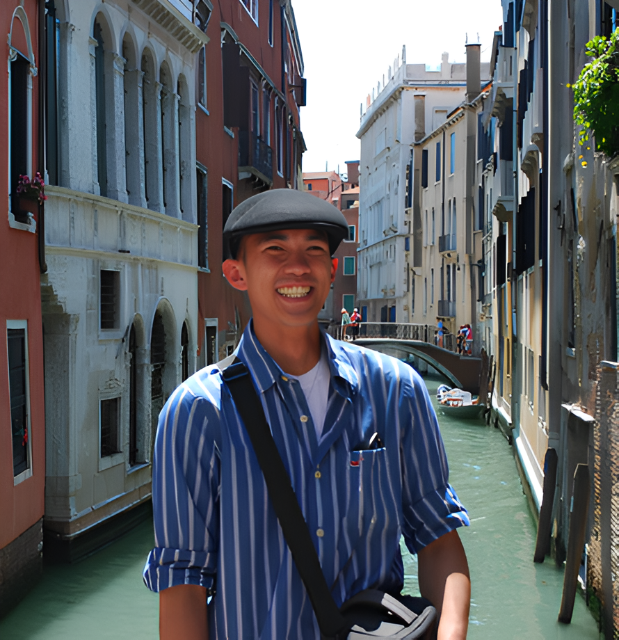 Smiling man in a flat cap and blue striped shirt standing beside a Venetian canal surrounded by historic buildings.