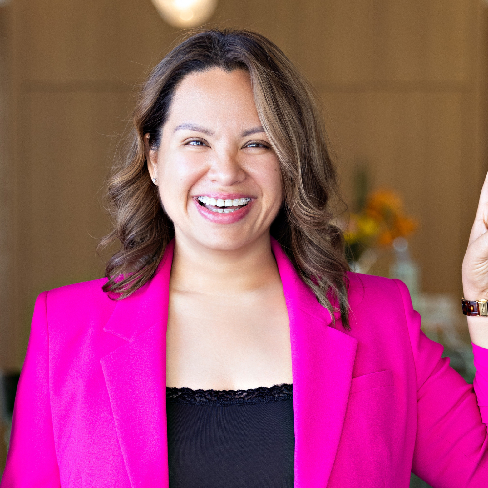 A woman in a pink blazer smiling with a raised hand in a bright indoor setting.