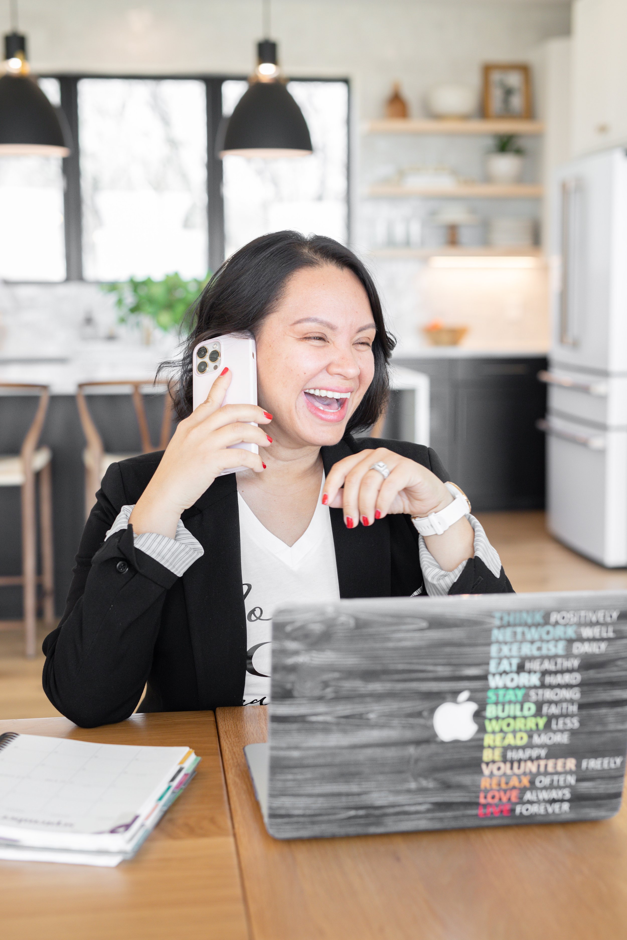 A woman with short black hair, dressed in a black blazer and white T-shirt, is laughing while talking on her cellphone. She sits at a wooden table with a laptop decorated with colorful words and a planner.