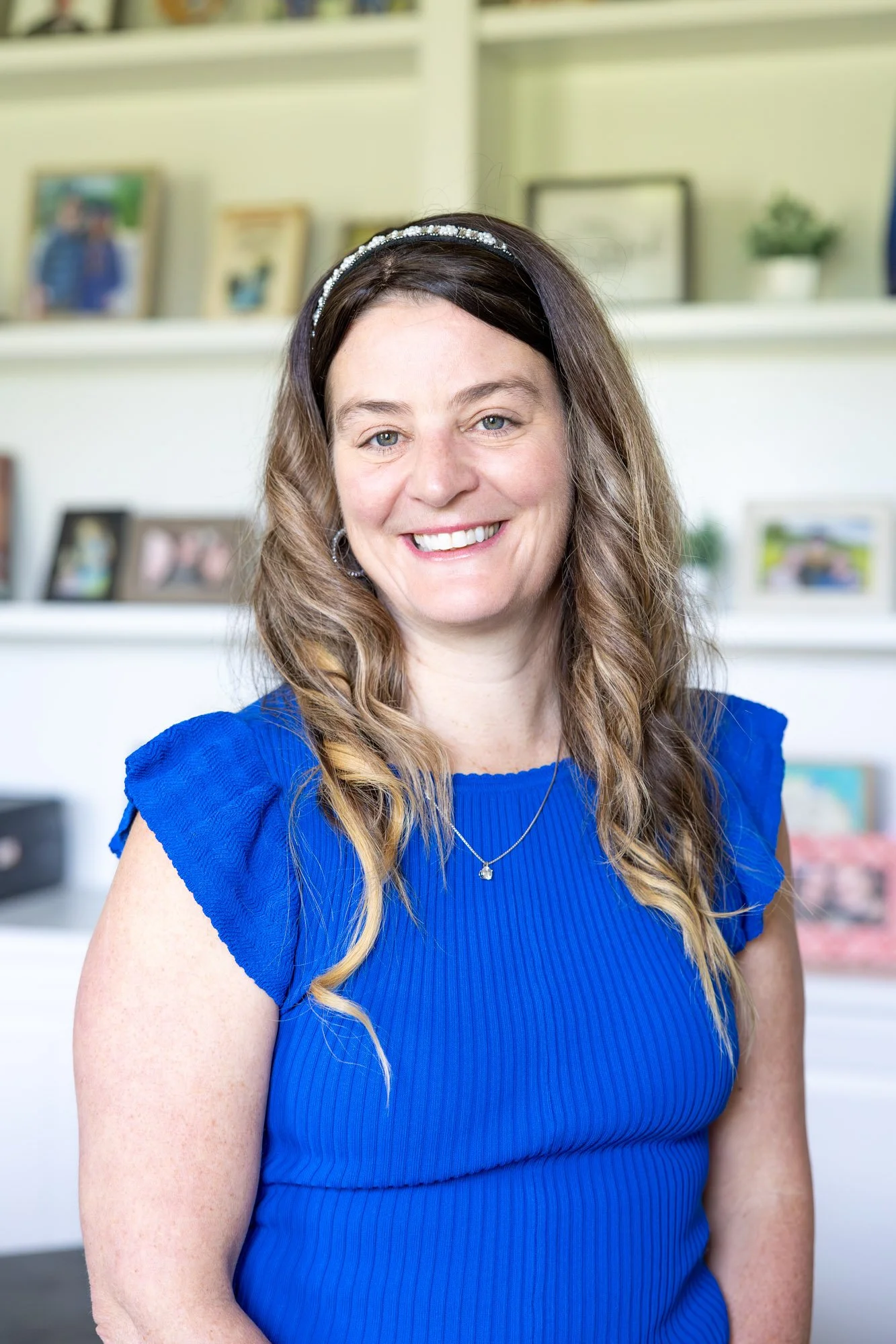 Portrait of a smiling woman with long, wavy hair, wearing a bright blue dress, a silver necklace, and a headband, standing in front of a bookshelf with framed photos and decorative items.