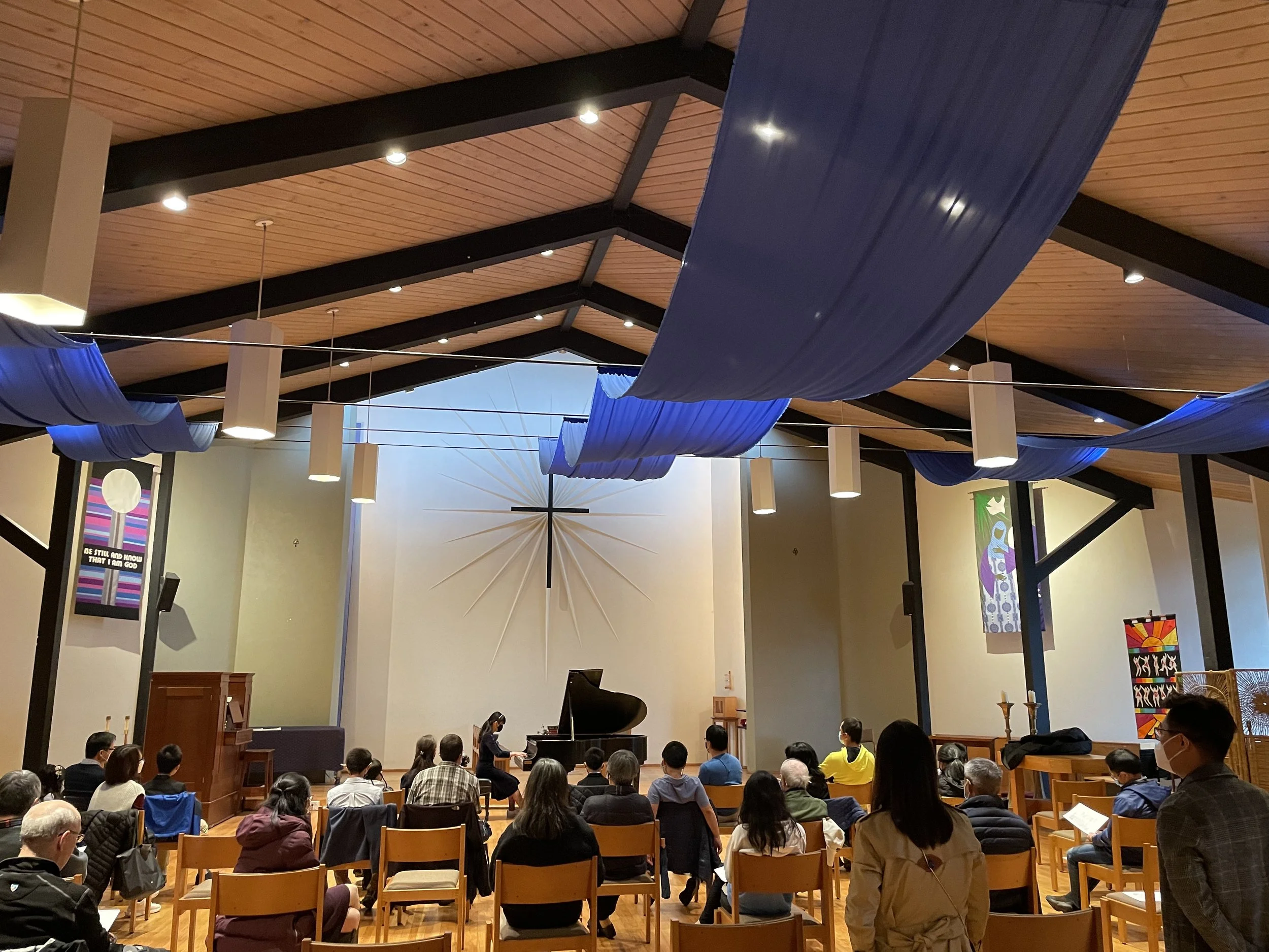 A diverse group of people sitting on chairs and listening to a woman playing the piano in a spacious, wood-paneled church or auditorium with blue fabric drapes hanging from the ceiling, colorful artwork on the walls, and a large star-like decoration on the wall behind the stage.