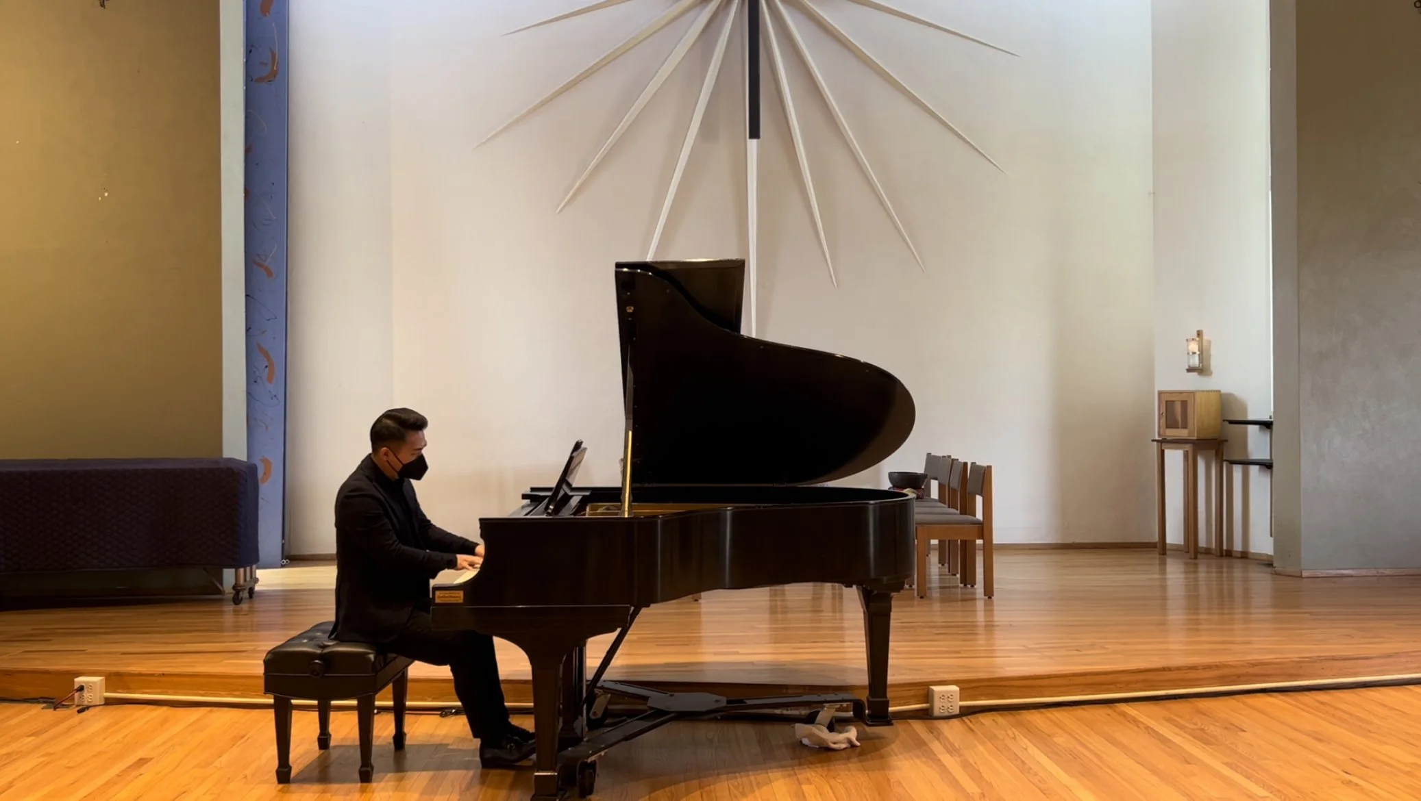 A pianist wearing a black suit and mask playing a grand piano on a stage with wooden flooring, behind a large decorative wall piece resembling a sunburst.