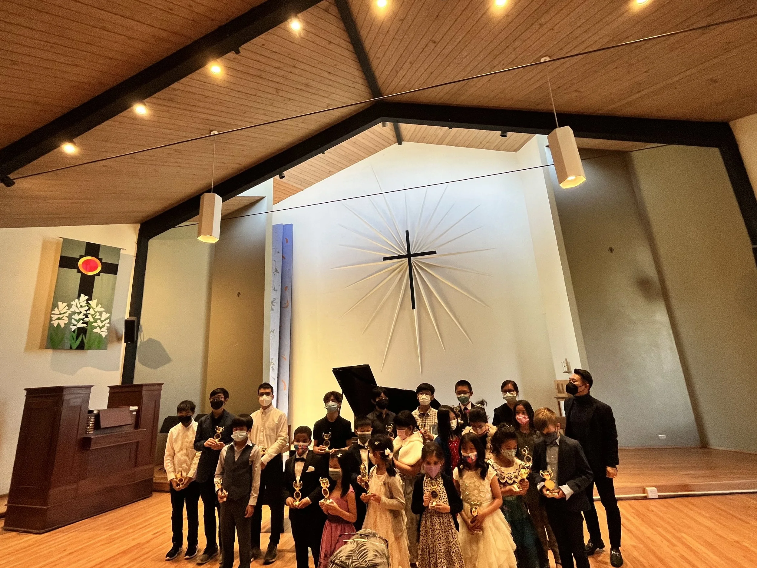 A group of children and teenagers wearing formal attire and face masks, holding trophies, posing for a photo in a wooden-ceiling hall during an award ceremony or concert.