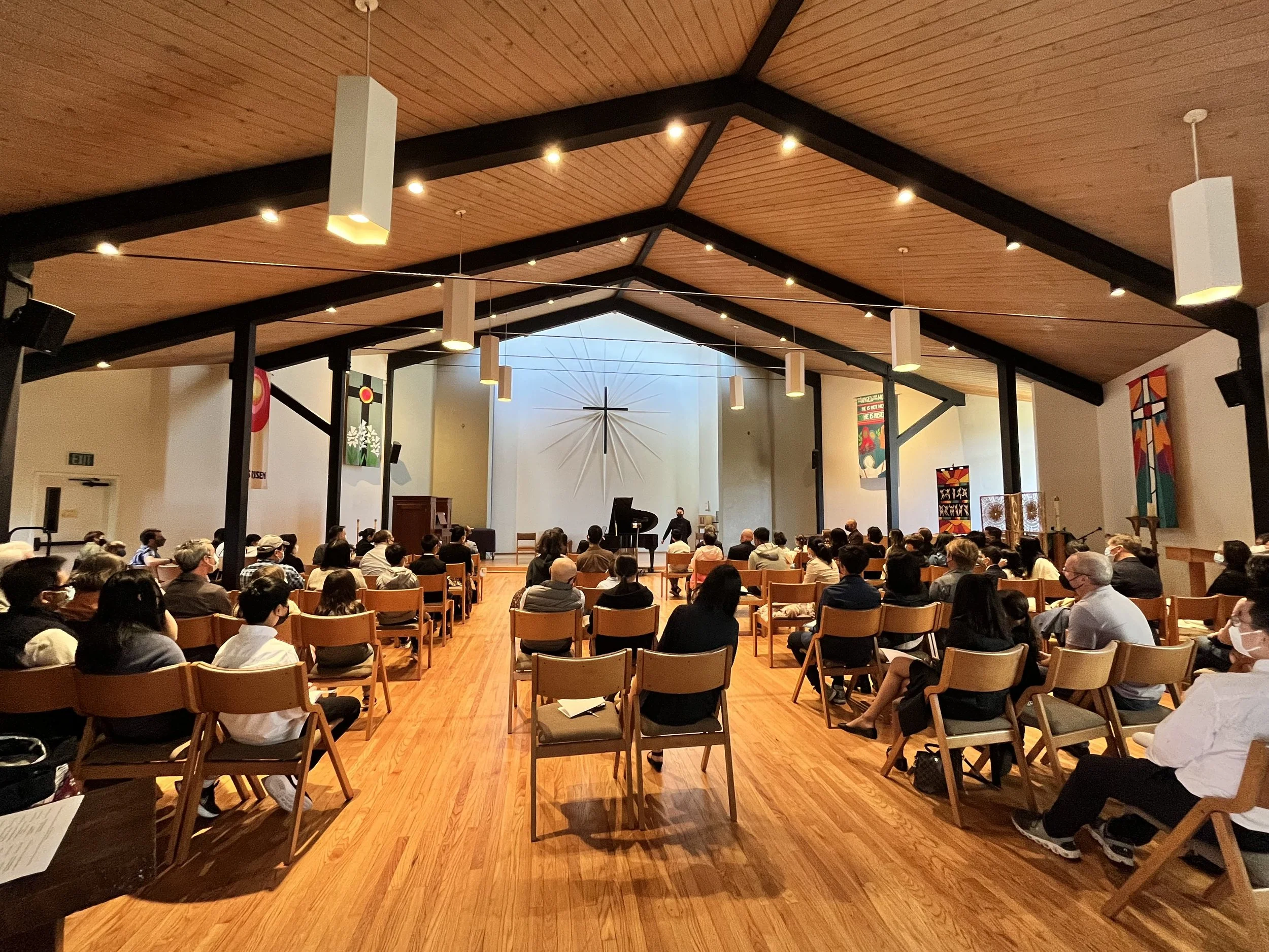 Inside a church with a congregation sitting on wooden chairs, listening to a performance at the front. The ceiling is wooden with hanging lights, and there is a cross on the wall behind the performers.