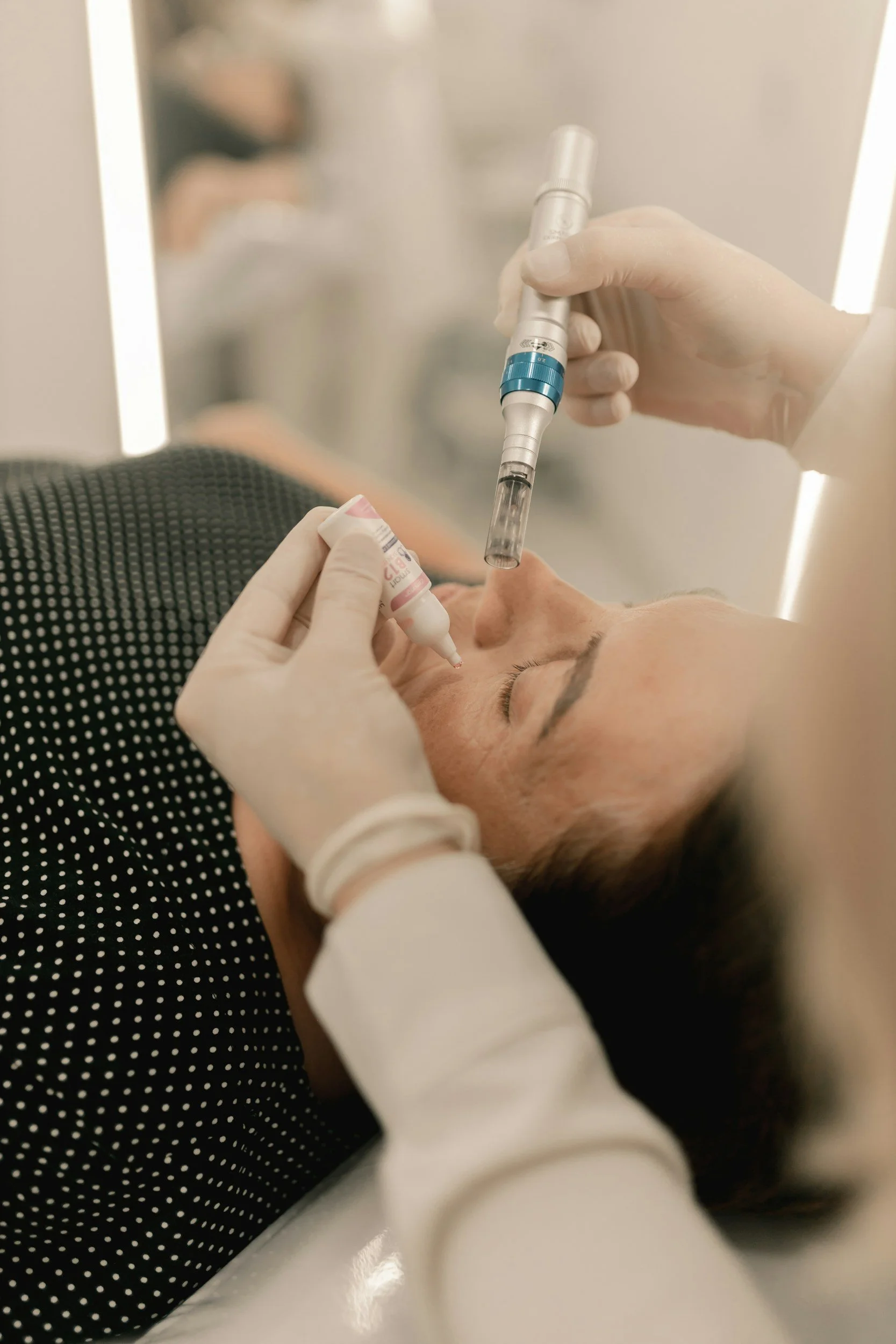 A medical professional appears to be administering a cosmetic or dermatological procedure on a woman lying down. The professional is holding a device and working on the woman's nose. The woman is wearing a black polka dot garment and has her eyes closed.