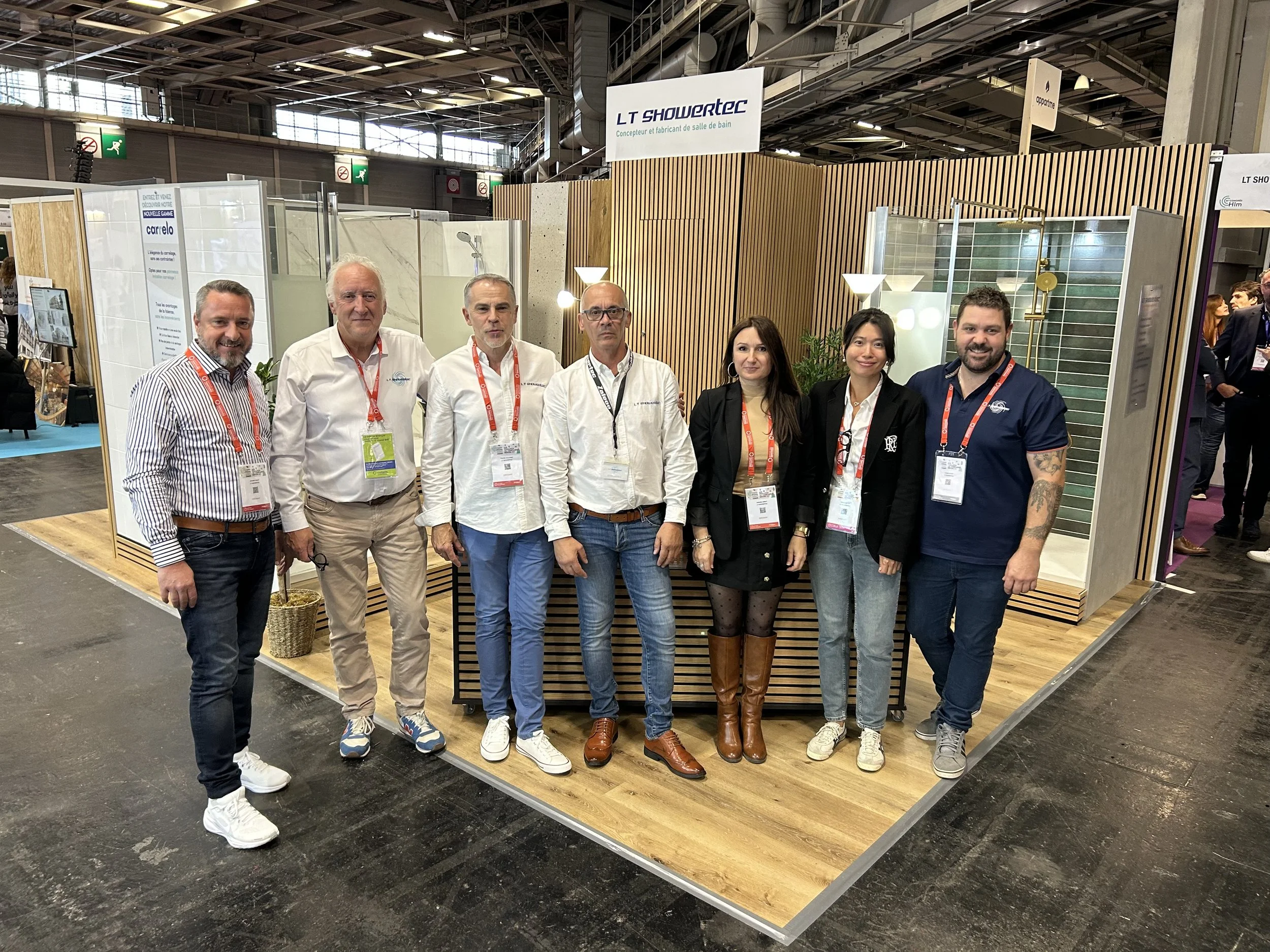 Groupe de sept personnes posant ensemble dans un stand d'exposition avec un panneau indiquant "LT Showertec". L'emplacement semble être une foire ou un salon professionnel, avec des stands et des visiteurs en arrière-plan.