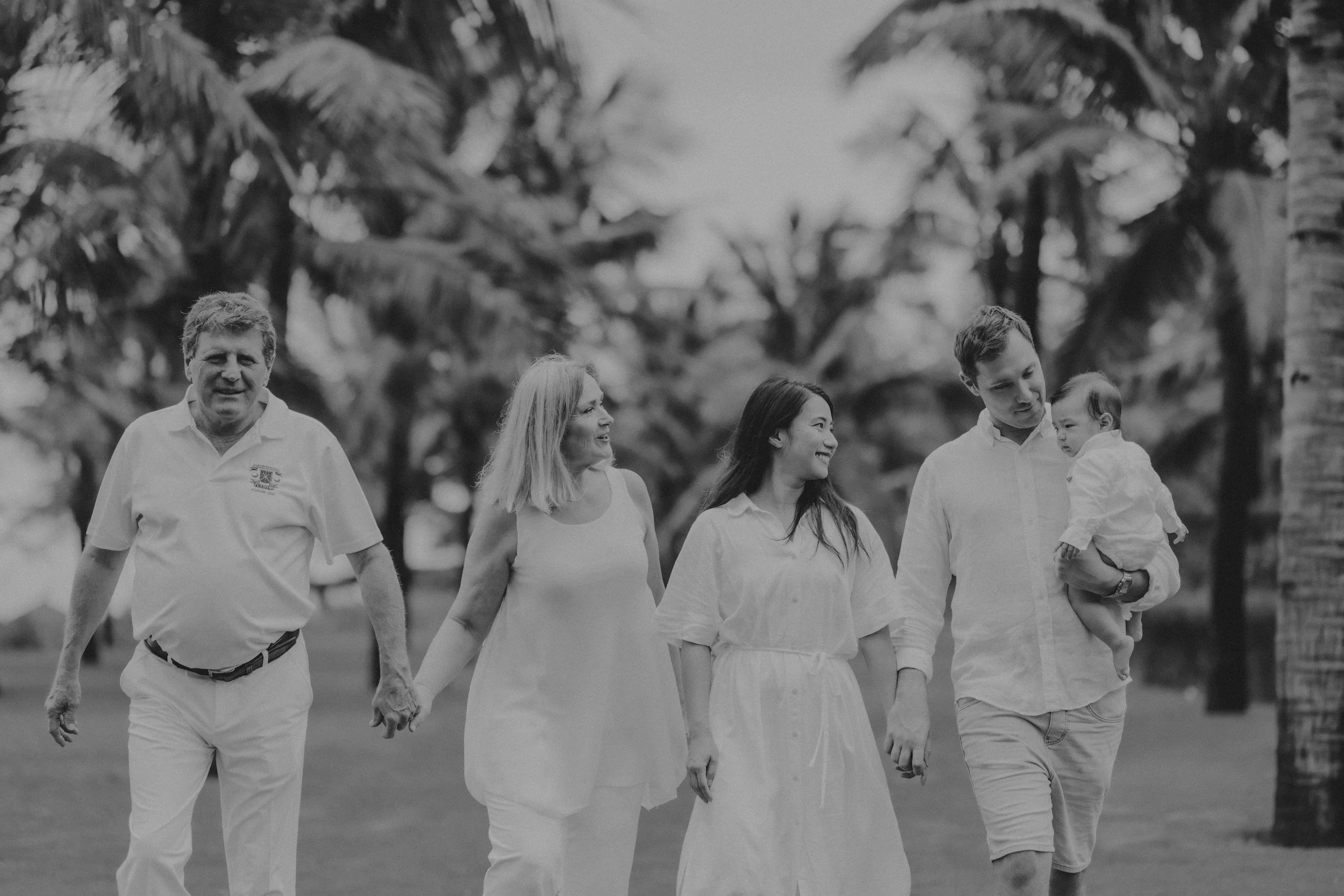 A family of five walking outdoors among palm trees, holding hands and smiling, with a man carrying a young child.