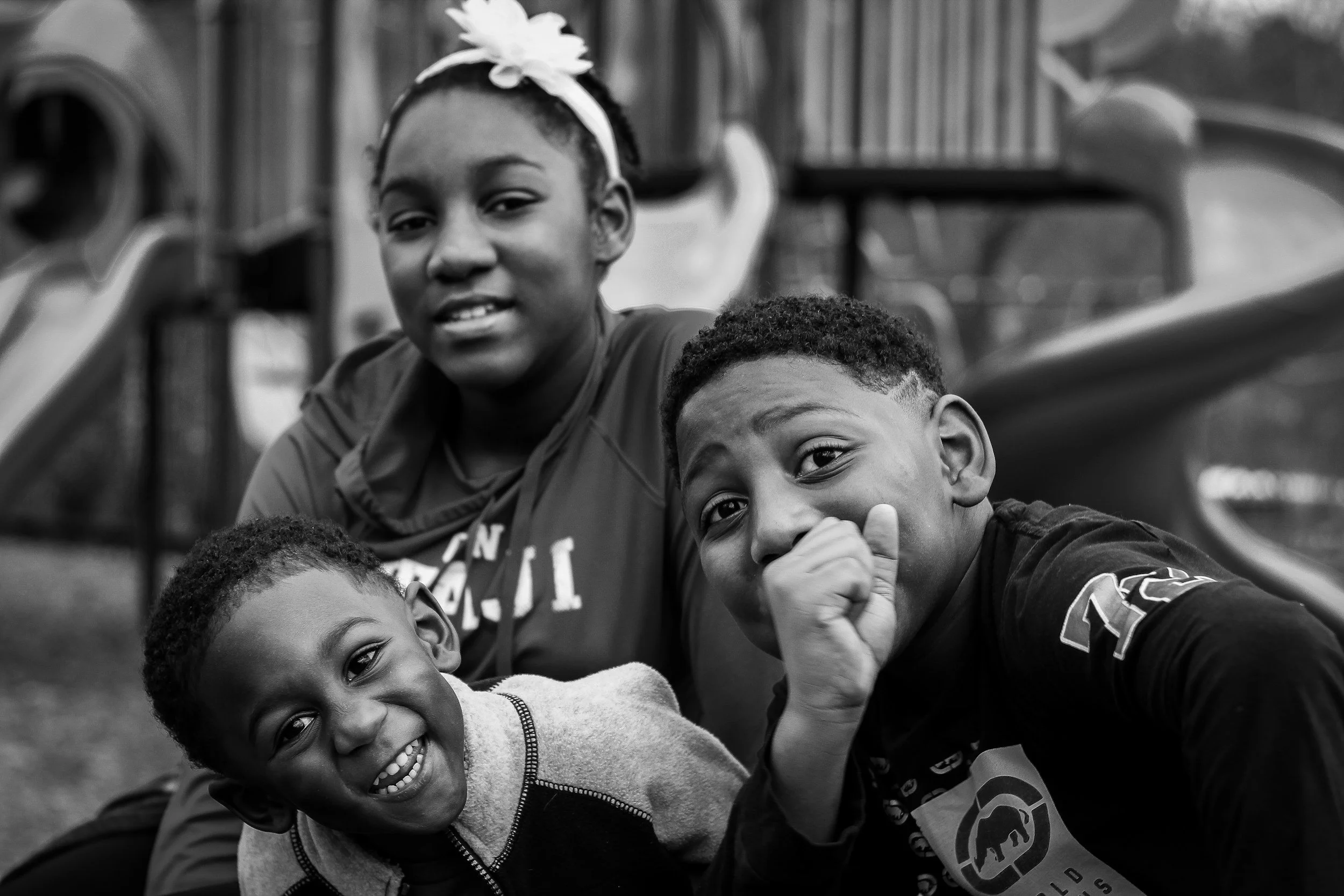 Four children playing outdoors at a playground, smiling and posing for the camera, with slides in the background.