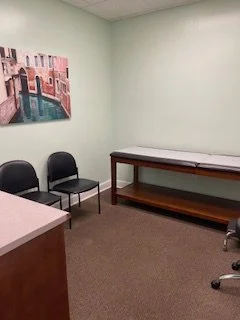 Empty office room with green walls, two black chairs, a wooden table, and a painting of a canal scene on the wall.