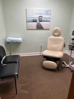 Medical examination room with a beige reclining chair, two black chairs, a small table, a painting of a pier with a lighthouse, and medical equipment.