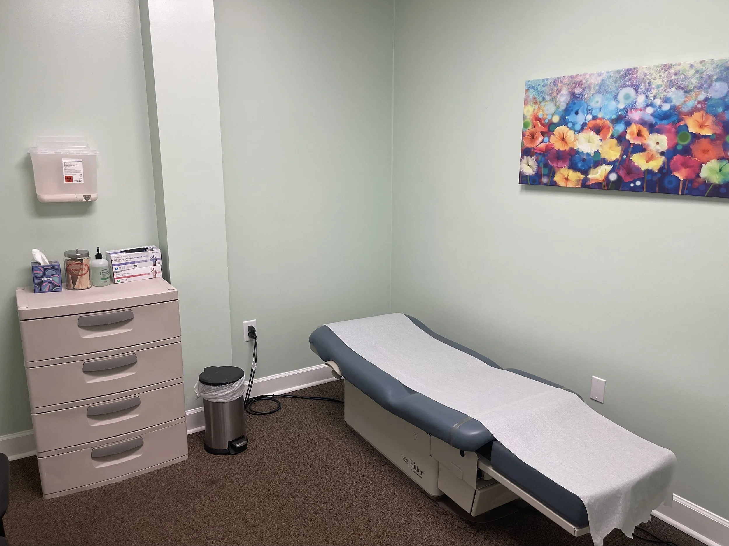 Medical examination room with a bed covered by a white sheet on one side, a beige drawer unit with medical supplies on top, a trash can, and a colorful floral painting on the wall.