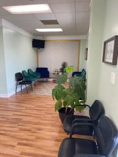 A waiting room with green chairs, black chairs, a small table, potted plants, framed pictures on the wall, and a ceiling-mounted speaker.