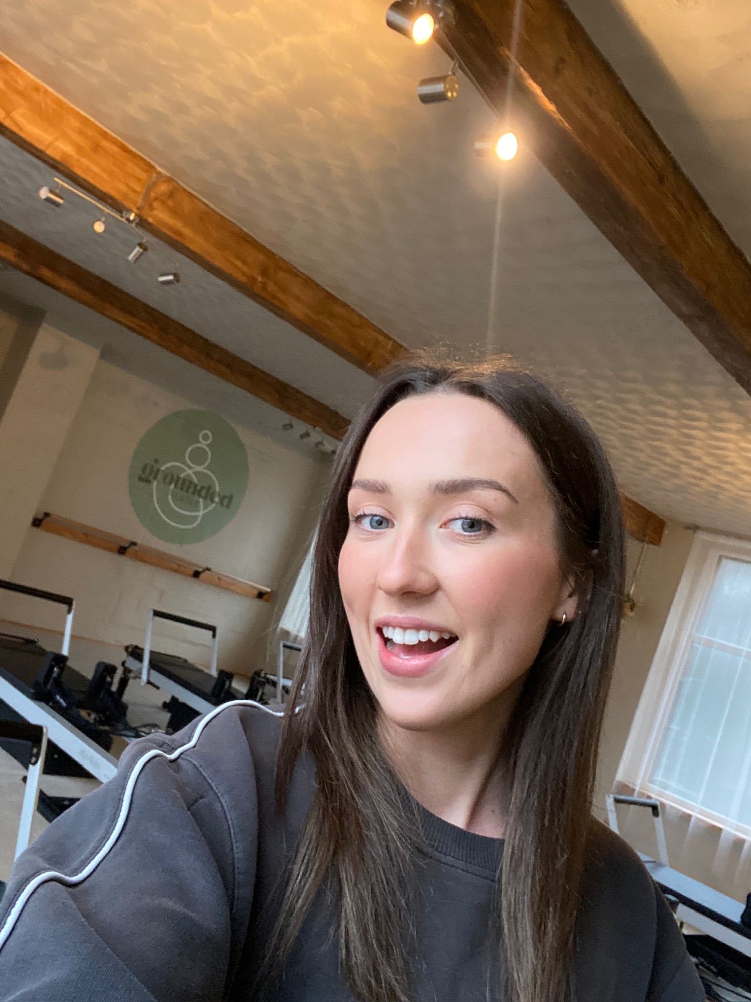 A woman with long brown hair and blue eyes taking a selfie in a fitness studio with wooden beams and workout equipment in the background.