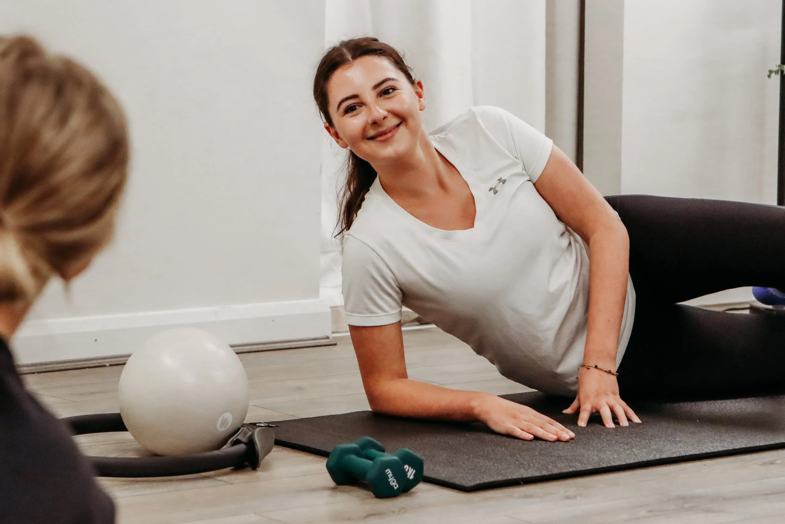 A woman with brown hair tied back, wearing a white Under Armour t-shirt, is doing a workout on a black mat inside a room with light-colored walls and blinds. She is smiling and supporting herself on her hands and knees. A white exercise ball and small green dumbbells are on the floor nearby.