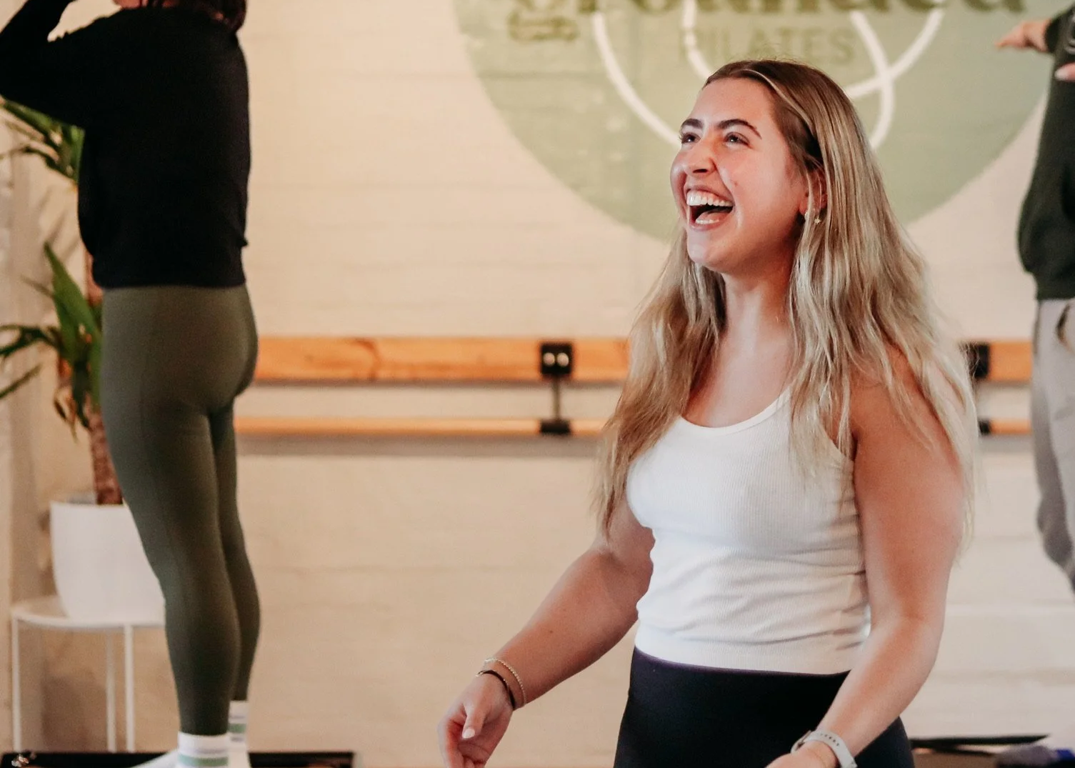A woman with long blonde hair, wearing a white tank top, smiling and laughing during a fitness class or workshop.