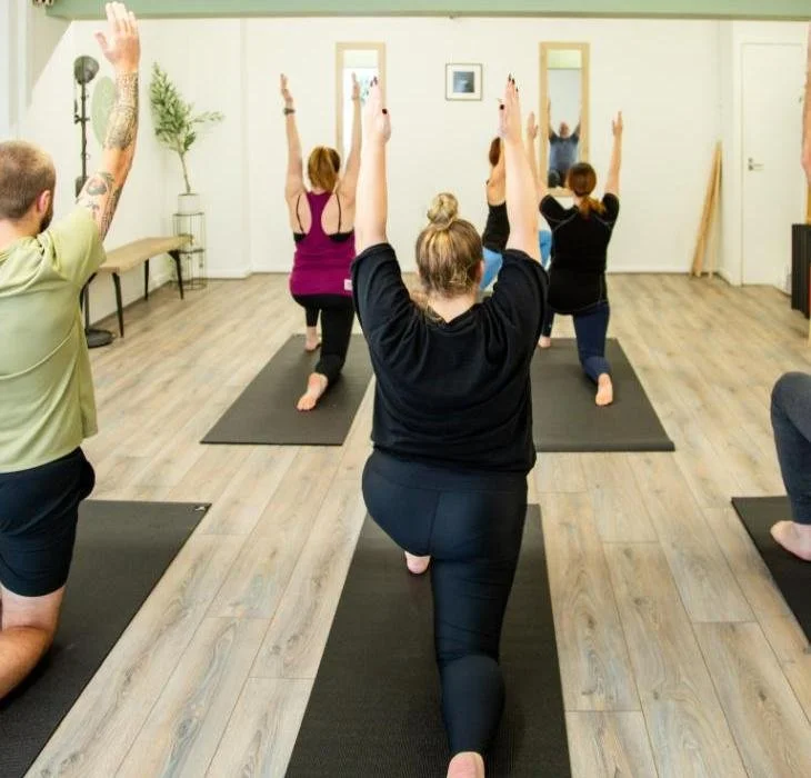 People practicing yoga in a spacious studio, kneeling on mats, raising their arms overhead in a back view.