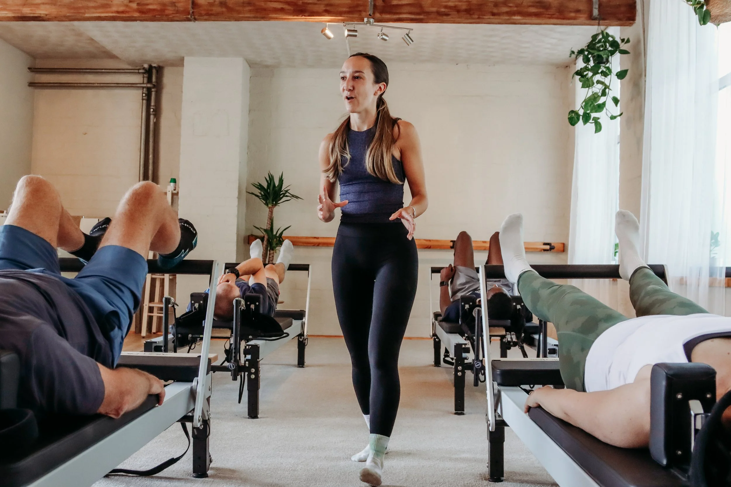 A woman leading a fitness class with people lying on exercise benches and raising their legs, in a well-lit room with plants and wooden ceiling beams.