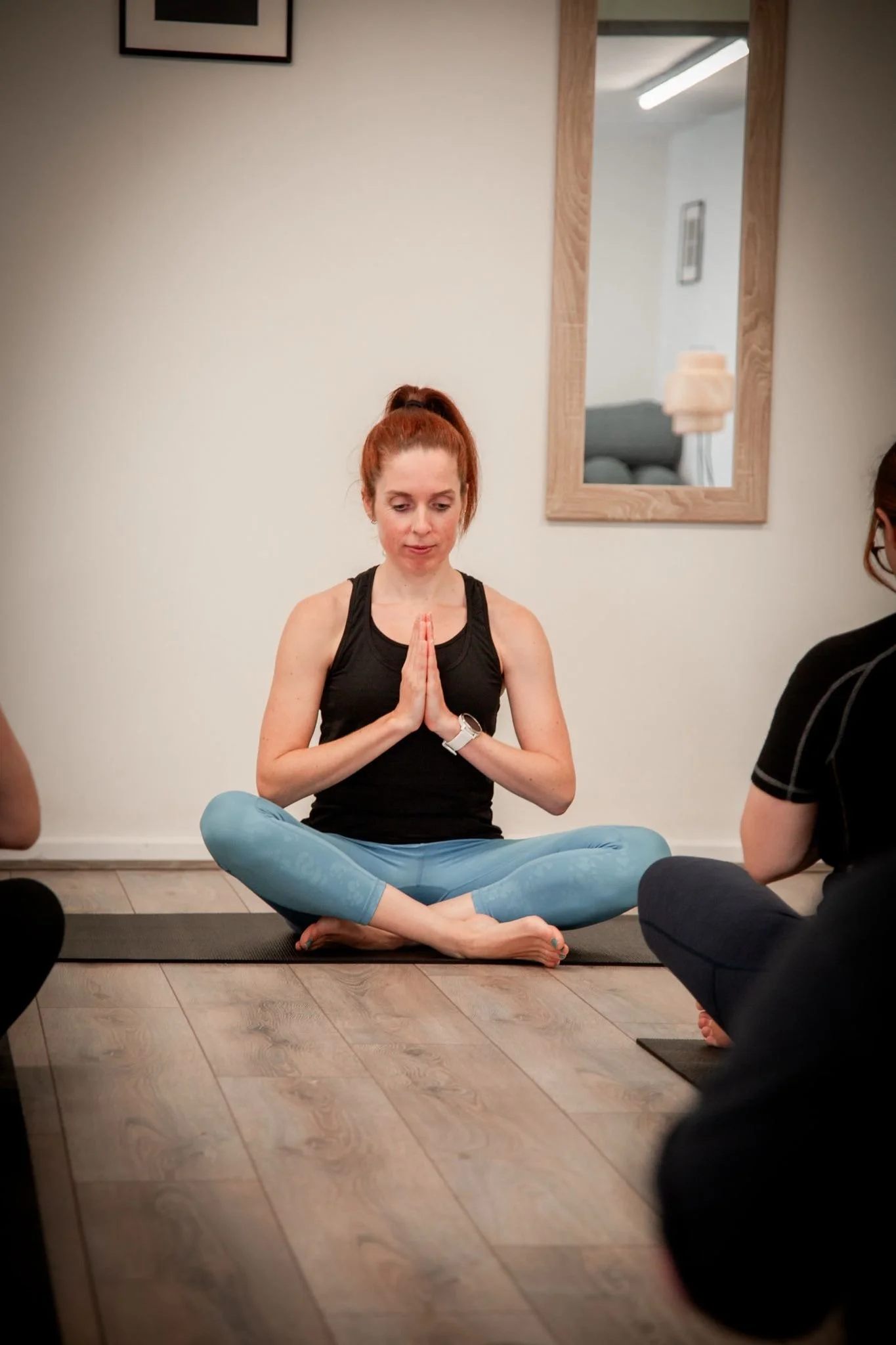 A woman in black tank top and blue leggings sitting cross-legged on a yoga mat with her hands in a prayer position, practicing yoga or meditation in a room with a large mirror on the wall.
