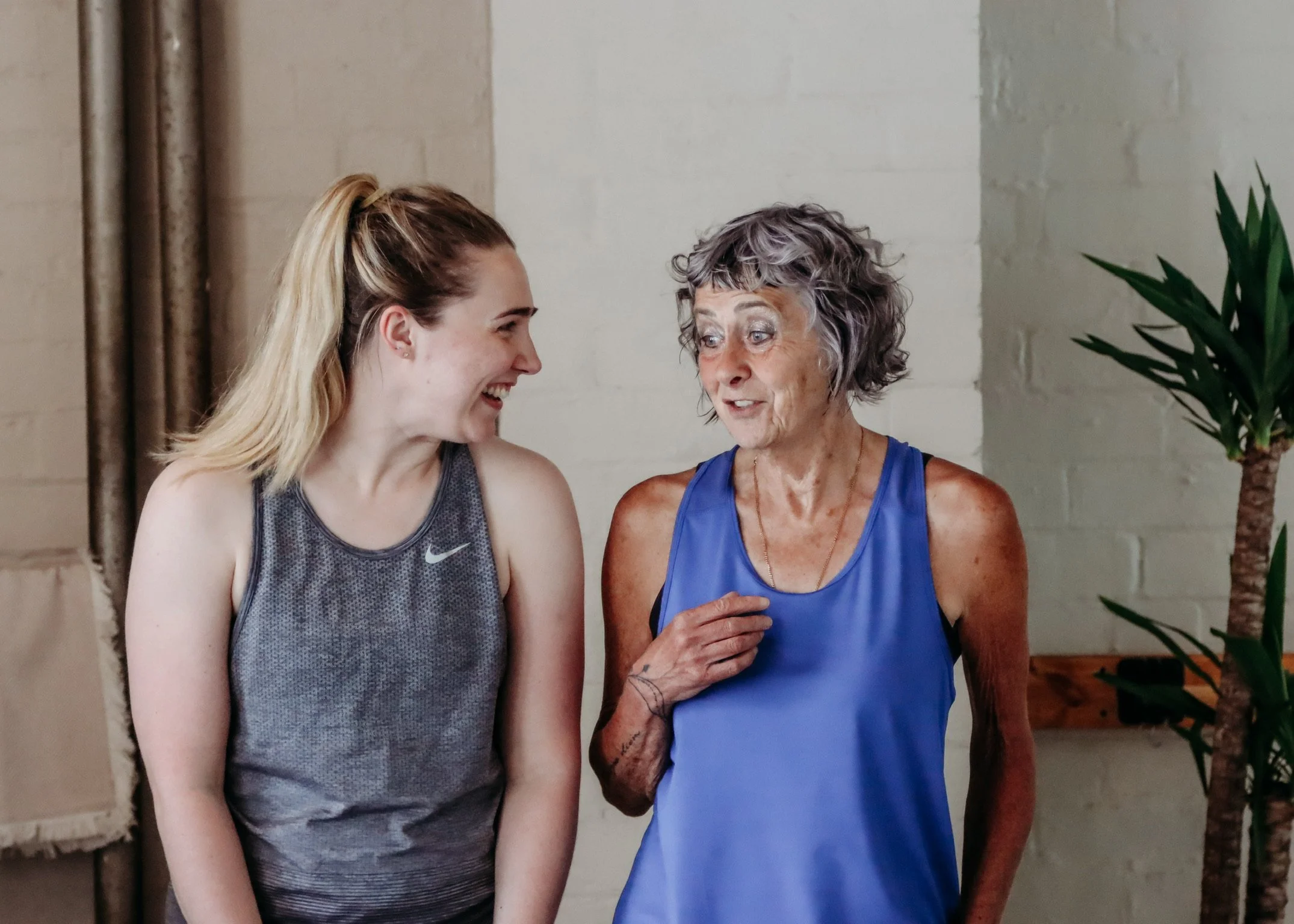 A young woman and an older woman are smiling and talking to each other indoors, with a plant visible on the right.