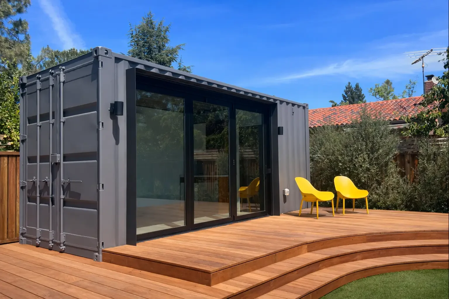A modern tiny home with a gray shipping container exterior and large glass sliding doors, situated on a wooden deck with two yellow chairs outside, in a backyard with trees and a house with a red tile roof under a blue sky.