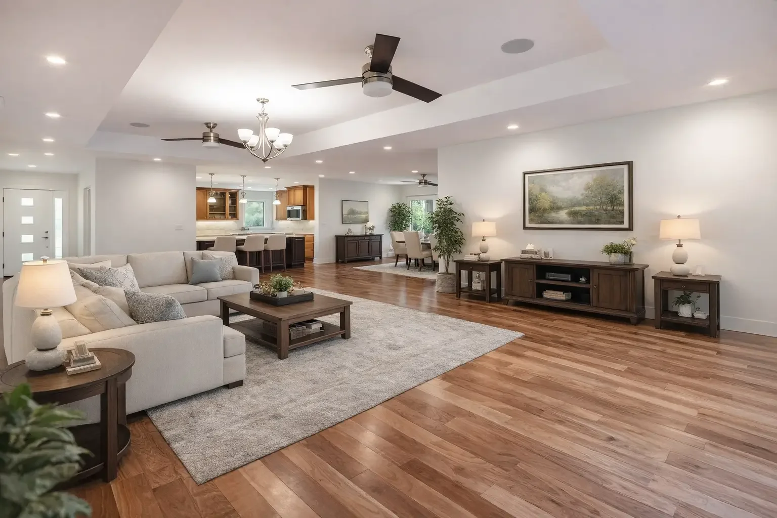 Spacious living room with white walls, hardwood floors, beige sofa with pillows, coffee table, side tables with lamps, landscape painting, TV stand, lamps, indoor plants, open kitchen with breakfast bar, kitchen cabinetry, dining area with table and chairs, ceiling fans, recessed lighting.