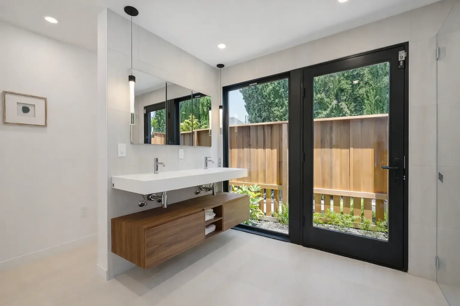 Modern bathroom with double sinks, large mirror, pendant lighting, and glass door leading outside to a small garden with a wooden fence.