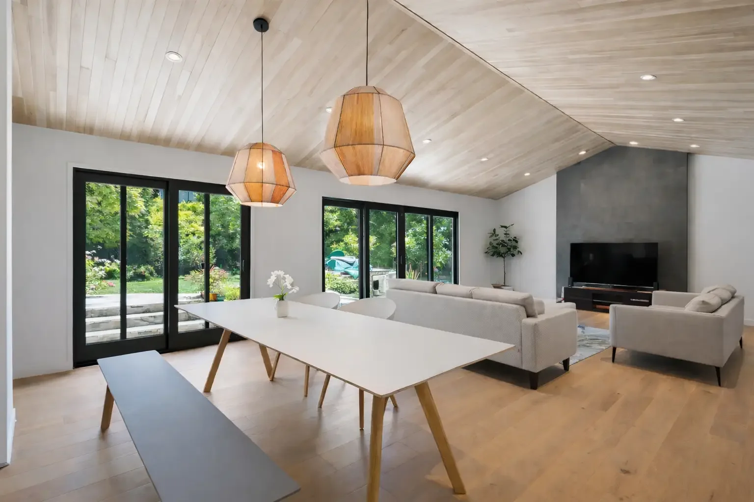 Modern living room with light wood ceiling, large windows with a garden view, a white dining table with wooden legs, two large pendant lights, and a seating area with a gray sofa, an armchair, a TV on a black stand, and a potted plant.