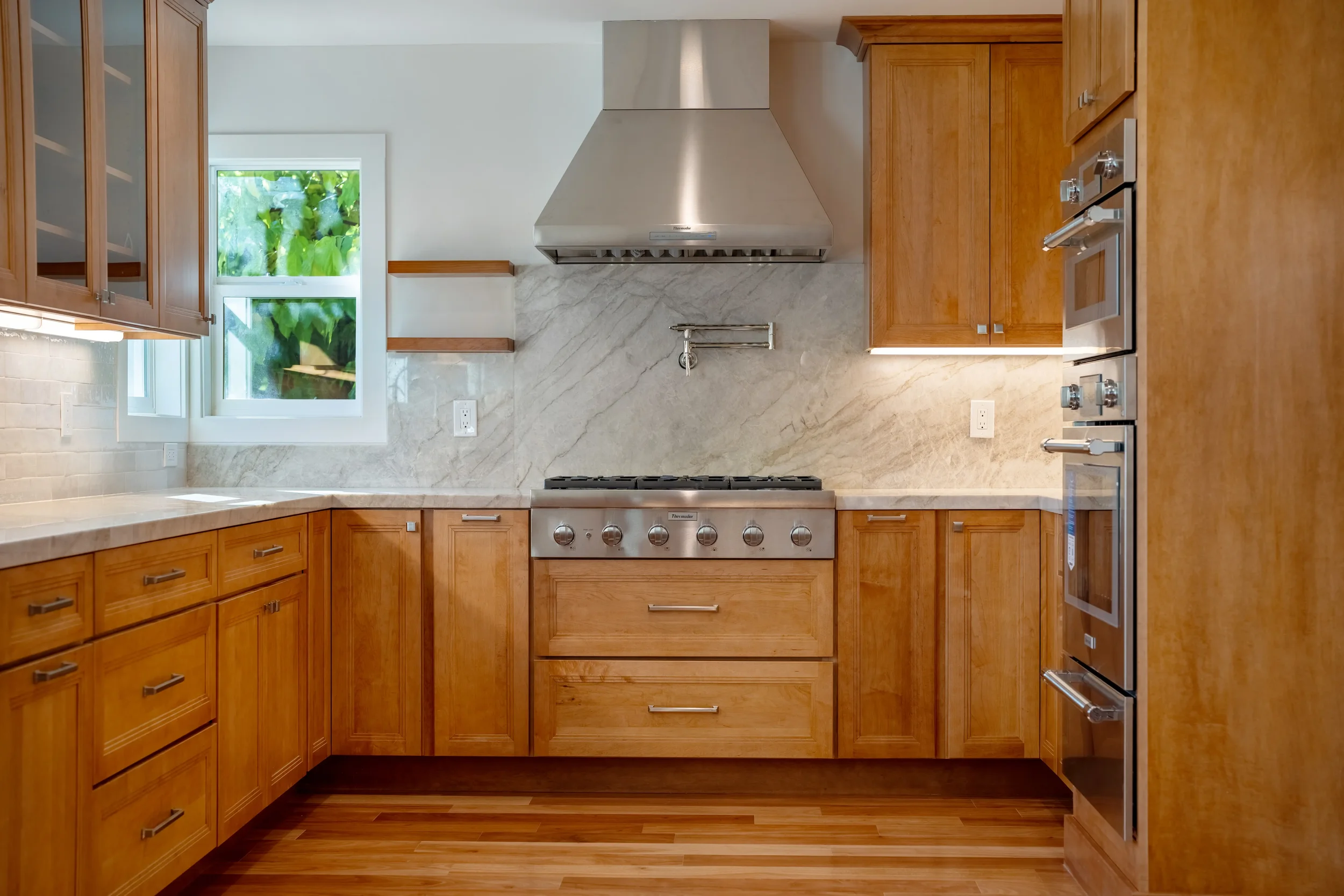 Kitchen with wooden cabinets, stainless steel appliances, marble backsplash, window with greenery outside, hardwood floor